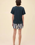 A person with short brown hair stands facing away against a plain beige background, wearing the DayDreamer John Lennon Peace Boyfriend Tee in lightweight cotton, black and white striped shorts, and black sandals.
