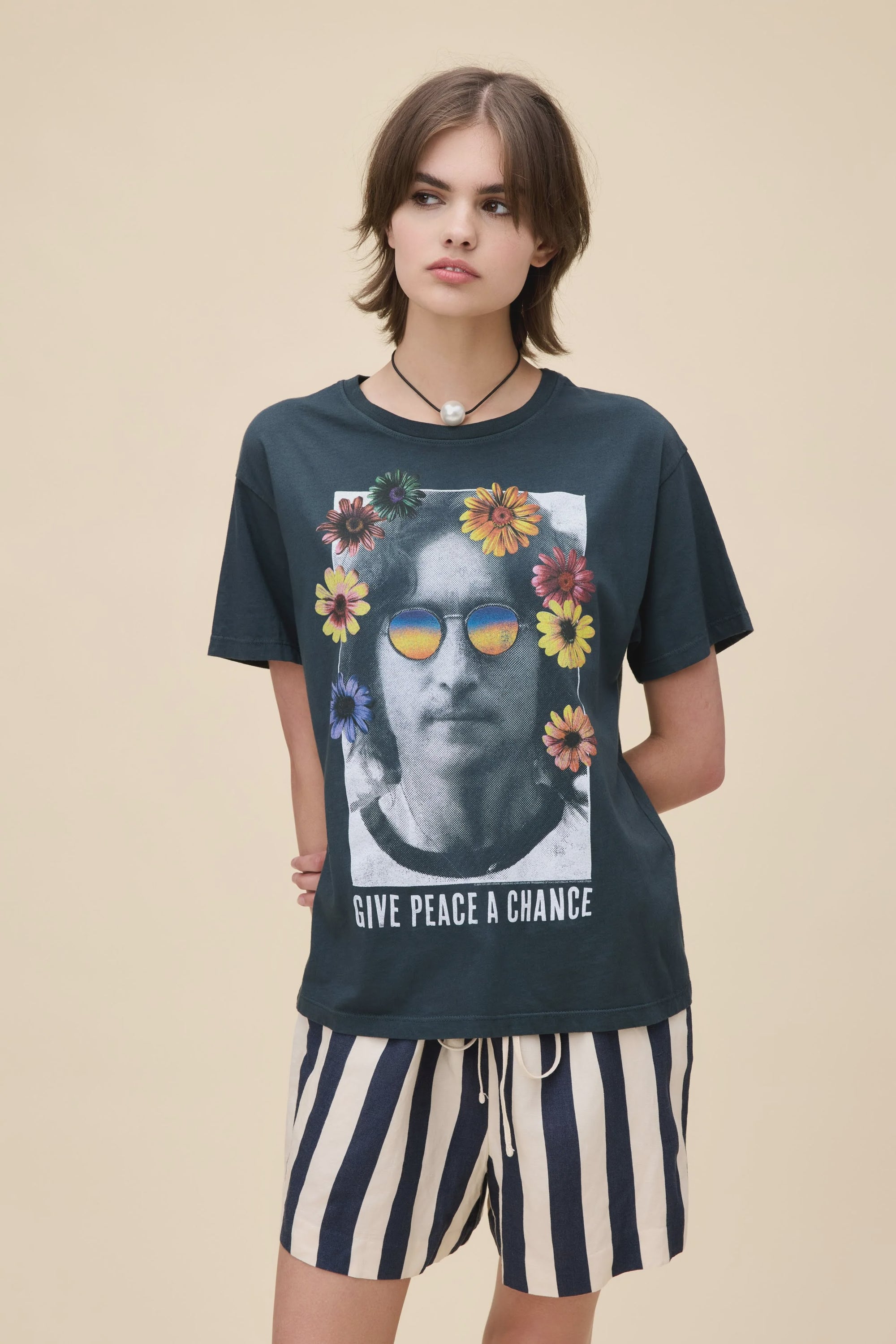 A young woman with short brown hair wears the DayDreamer "John Lennon Peace" lightweight cotton Boyfriend Tee, featuring John Lennon's face with colorful flowers and sunglasses, plus the text "GIVE PEACE A CHANCE," paired with striped shorts.