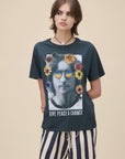 A young woman with short brown hair wears the DayDreamer "John Lennon Peace" lightweight cotton Boyfriend Tee, featuring John Lennon's face with colorful flowers and sunglasses, plus the text "GIVE PEACE A CHANCE," paired with striped shorts.