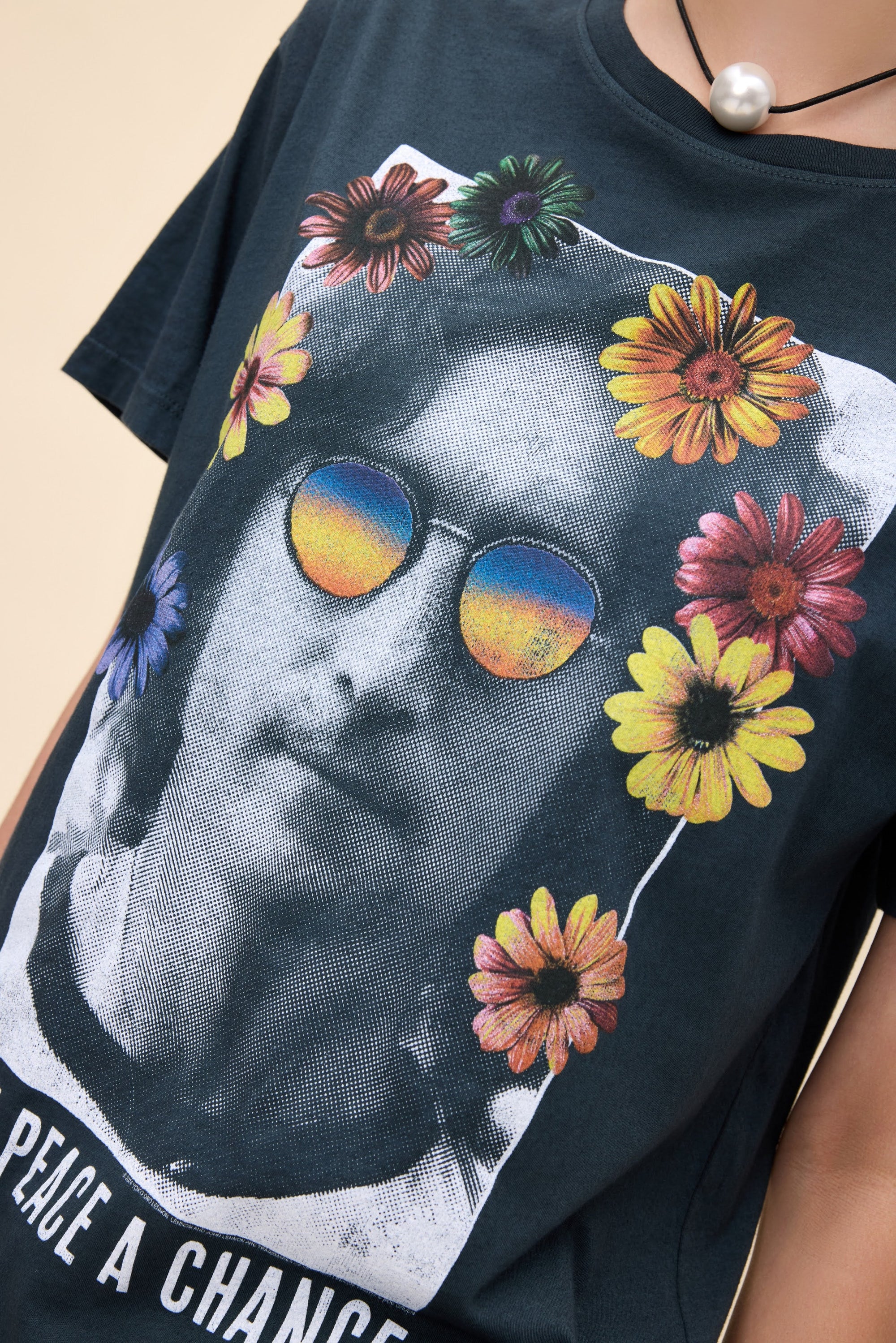 A person wears the DayDreamer John Lennon Peace tee, a lightweight cotton top featuring Lennon’s black-and-white portrait with colorful flower and rainbow sunglasses graphics, "PEACE A CHANCE" text, and completes the look with a statement pearl necklace.