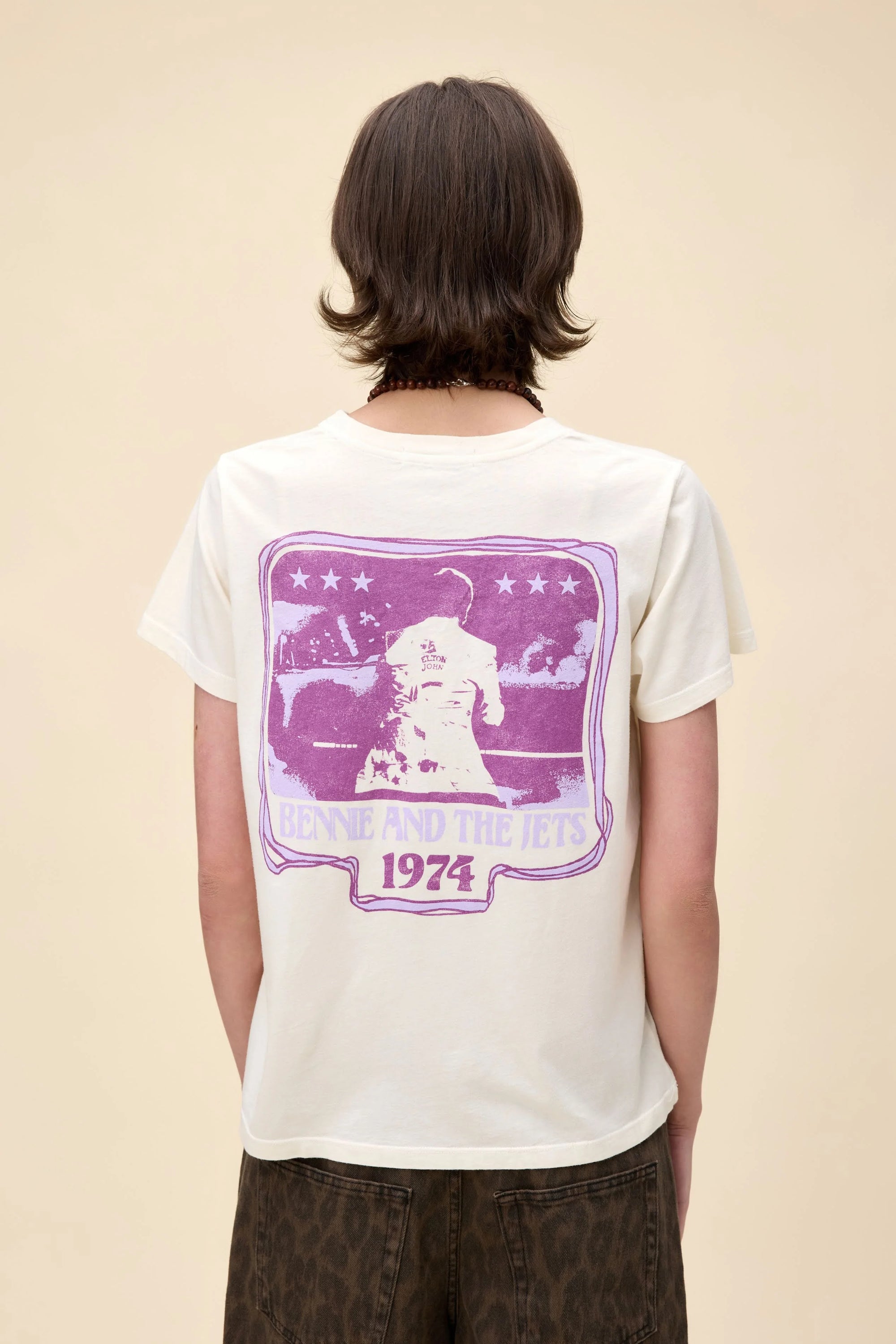 A person with short dark hair wears a white DayDreamer Elton John Bennie/Jets tee, featuring a purple "BENNIE AND THE JETS 1974" graphic and Elton John performing with stars on the back, standing before a plain beige background.