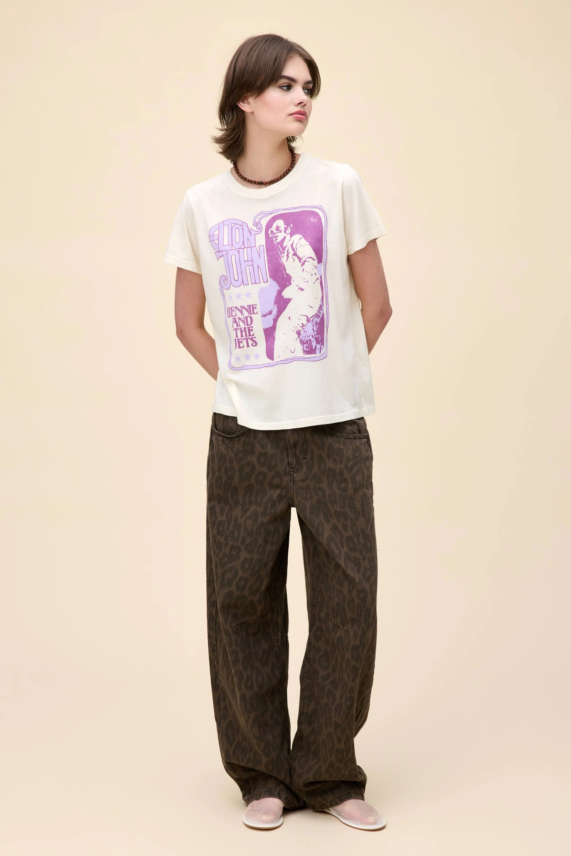 A person stands against a beige background, wearing the DayDreamer Elton John Bennie/Jets cream tee with a purple Elton John graphic, loose brown leopard print pants, and light shoes. Their hands are behind their back as they look to the side.
