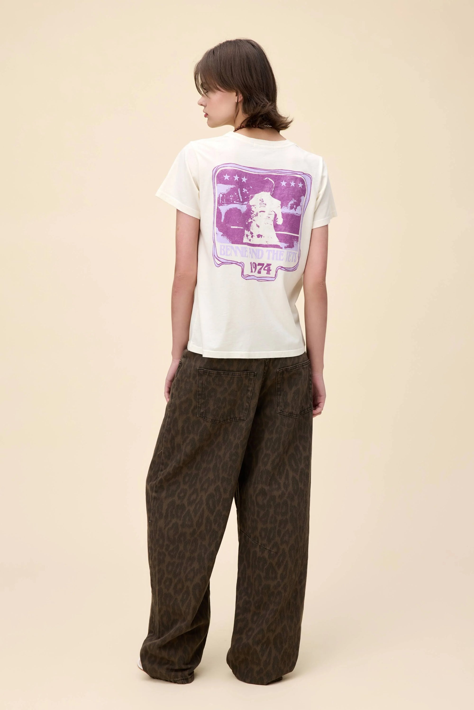 A person stands with their back to the camera, wearing the DayDreamer Elton John Bennie/Jets tee featuring a purple graphic and text, paired with loose dark brown leopard-print pants, set against a plain beige background.