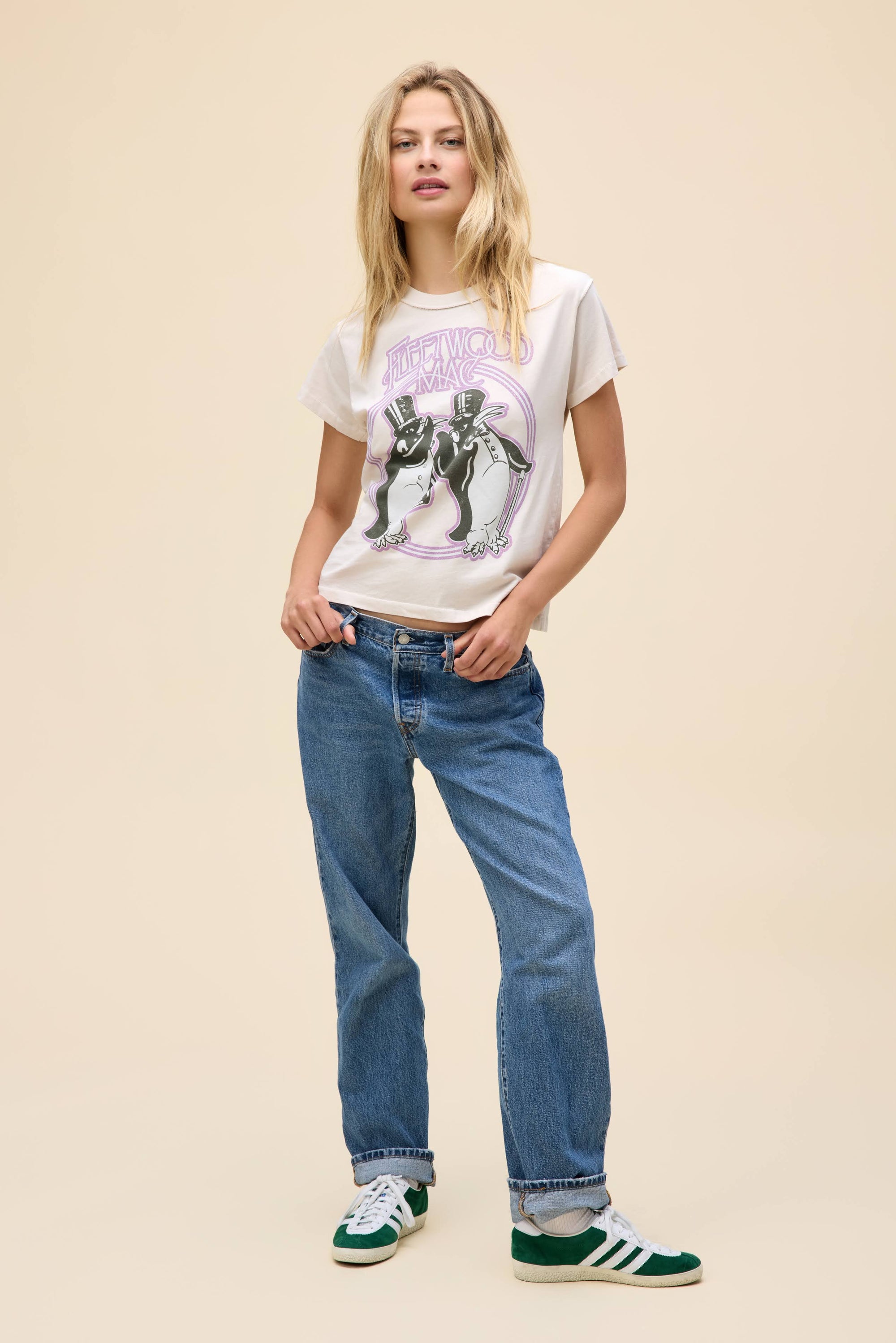 A woman poses against a beige background in DayDreamer’s Fleetwood Mac Penguins tee, blue jeans, and green Adidas sneakers—her relaxed style and blonde hair make this look perfect for any Fleetwood Mac fan.