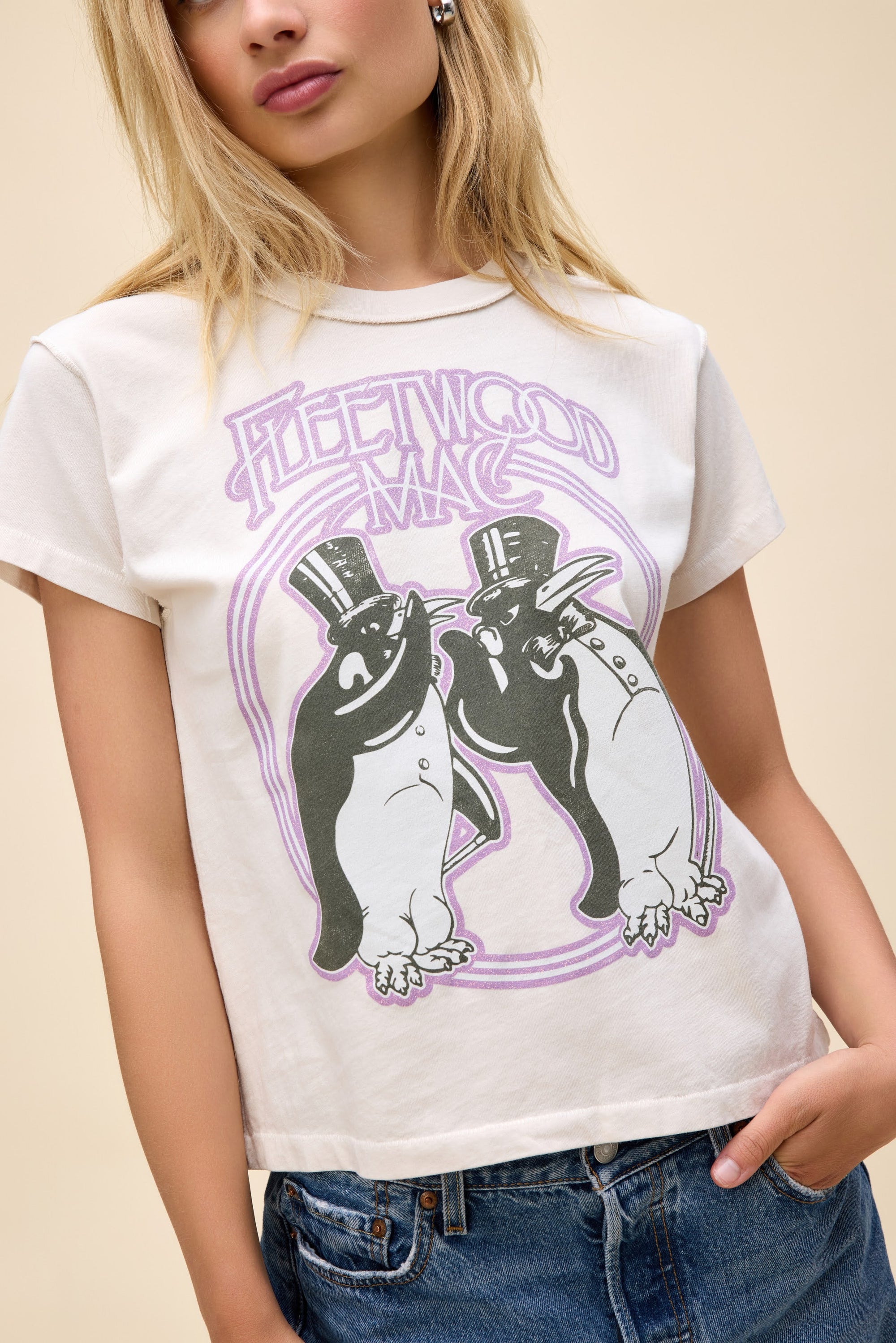 A woman with light hair and blue jeans models the DayDreamer Fleetwood Mac Penguins tee, a vintage-inspired white shirt featuring two top-hatted penguins, standing against a plain beige background.
