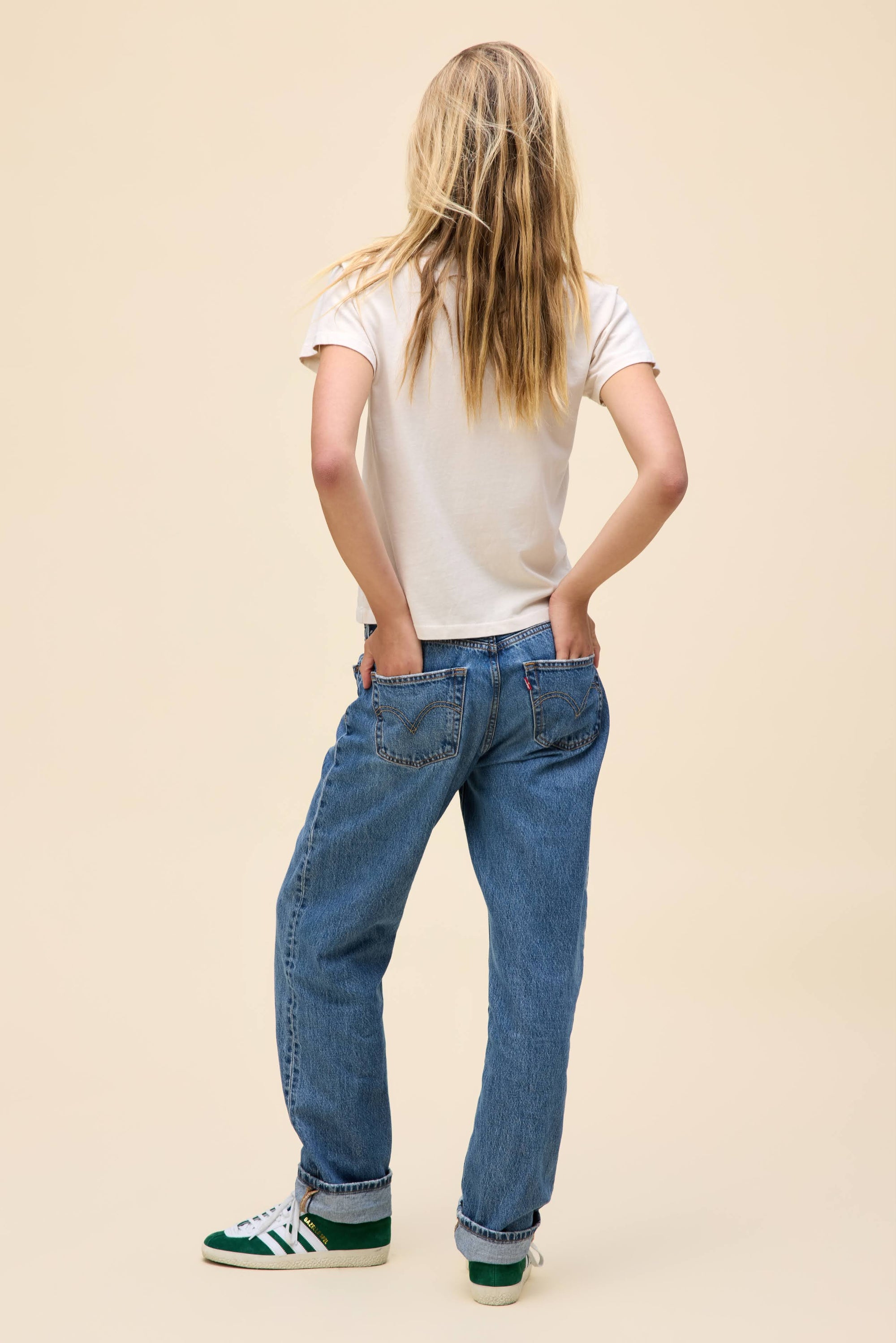 A person with long blonde hair, wearing a DayDreamer Fleetwood Mac Penguins Reverse GF Tee, blue jeans, and green sneakers stands facing away with hands in their back pockets against a plain beige background.