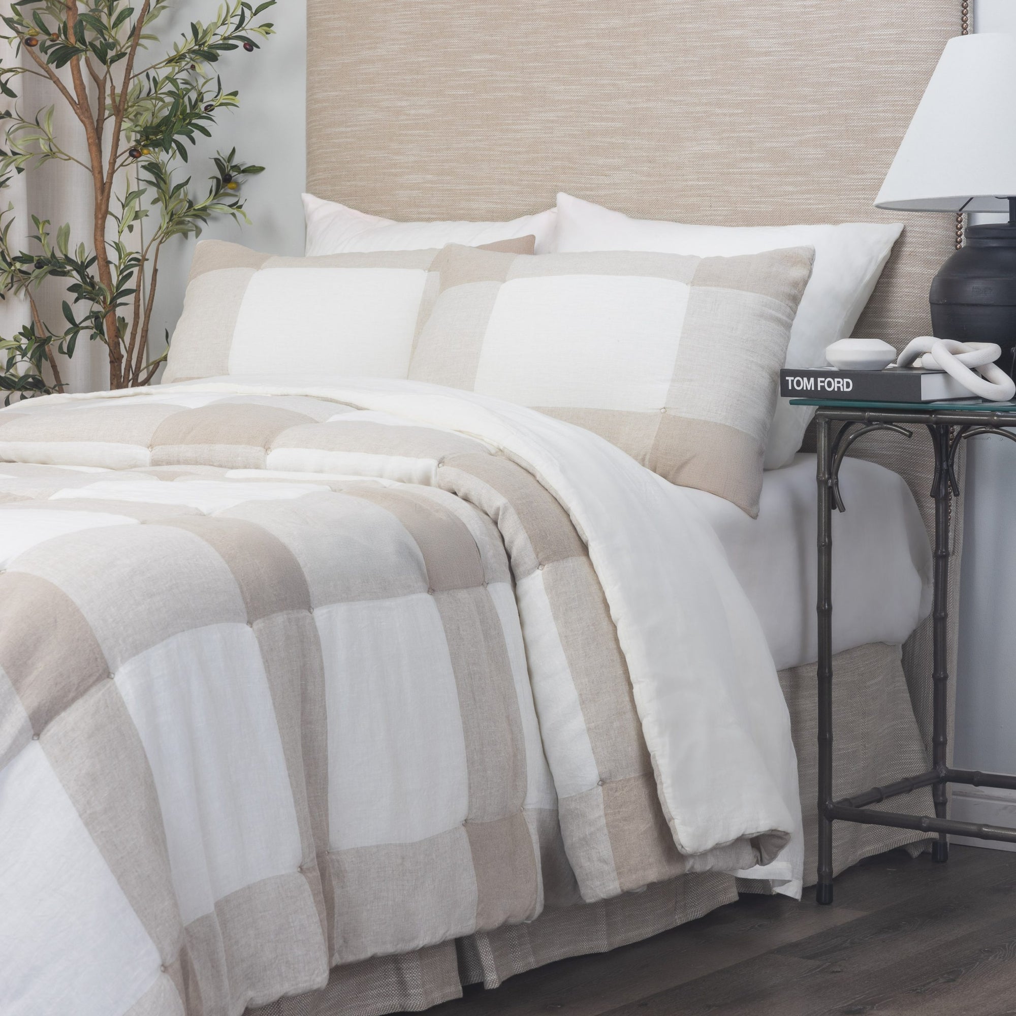 A neatly made bed features the Amity Home Chalmers Quilt Nat K in beige and white buffalo check, with two large pillows and a light wood headboard. A nightstand holds books, a decorative item, and a potted plant sits beside the bed.