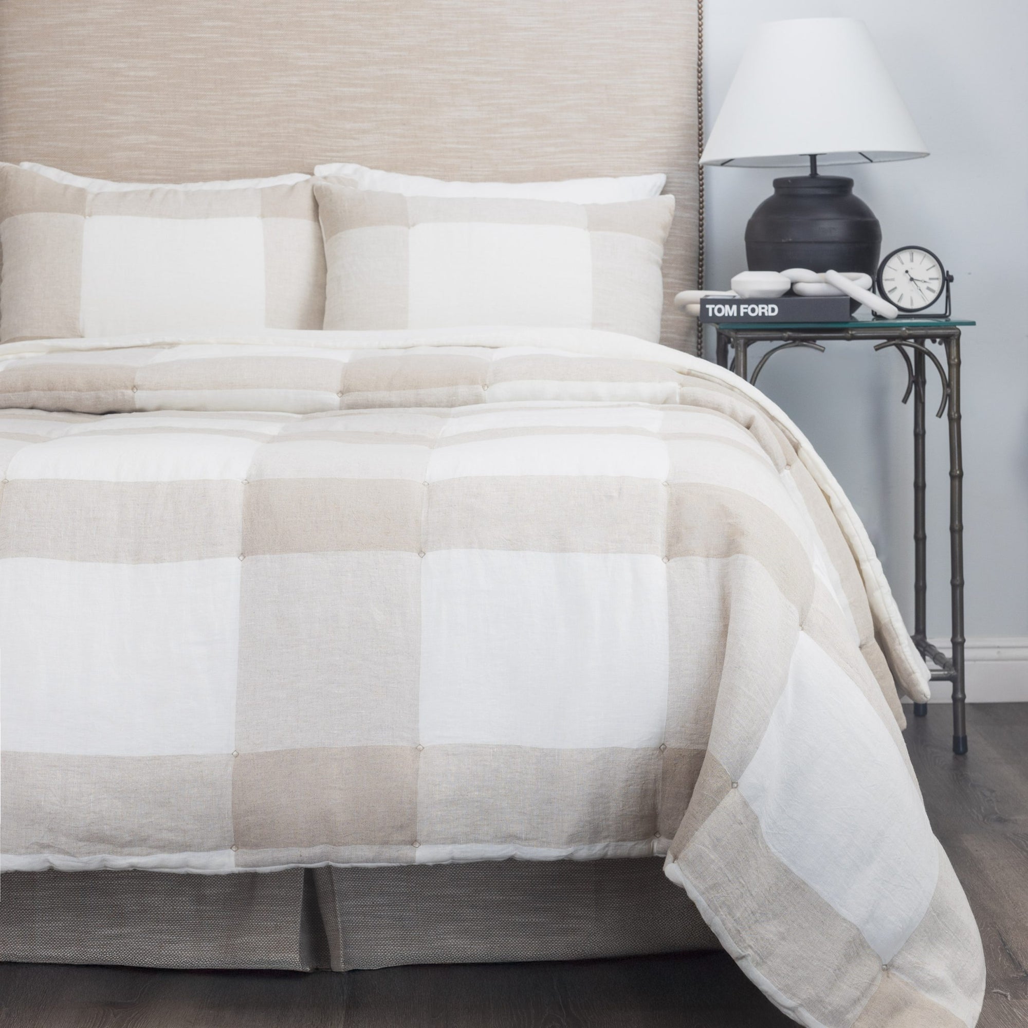 A neatly made bed with the Amity Home Chalmers Quilt Nat K and matching pillows sits beside a small table holding a black lamp, a clock, a “Tom Ford” book, and a candle.