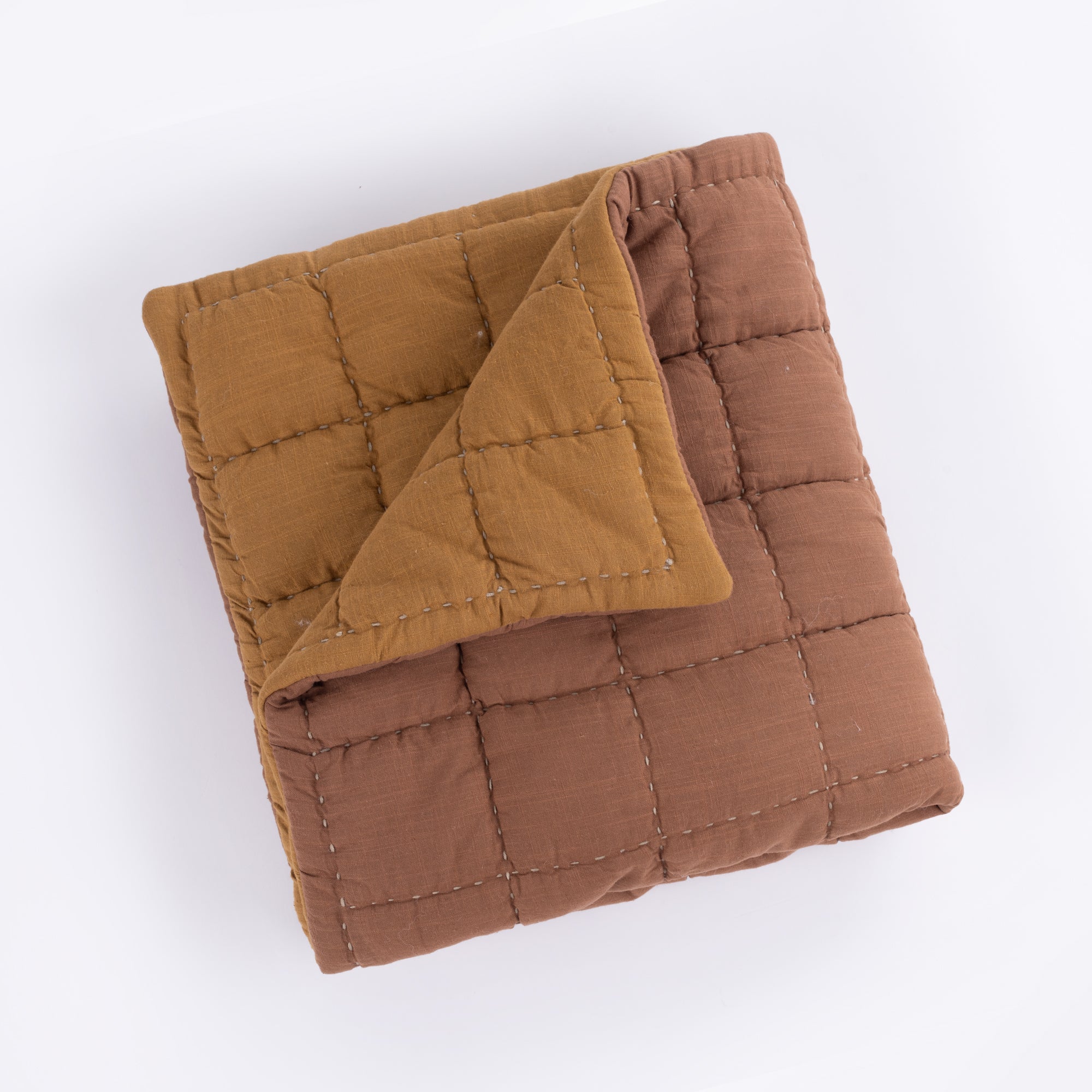 The Spencer Quilt by Amity Home is a reversible, folded, quilted blanket with a grid pattern in brown and mustard yellow slub cotton. Visible square stitching stands out, and the quilt is displayed against a white background.