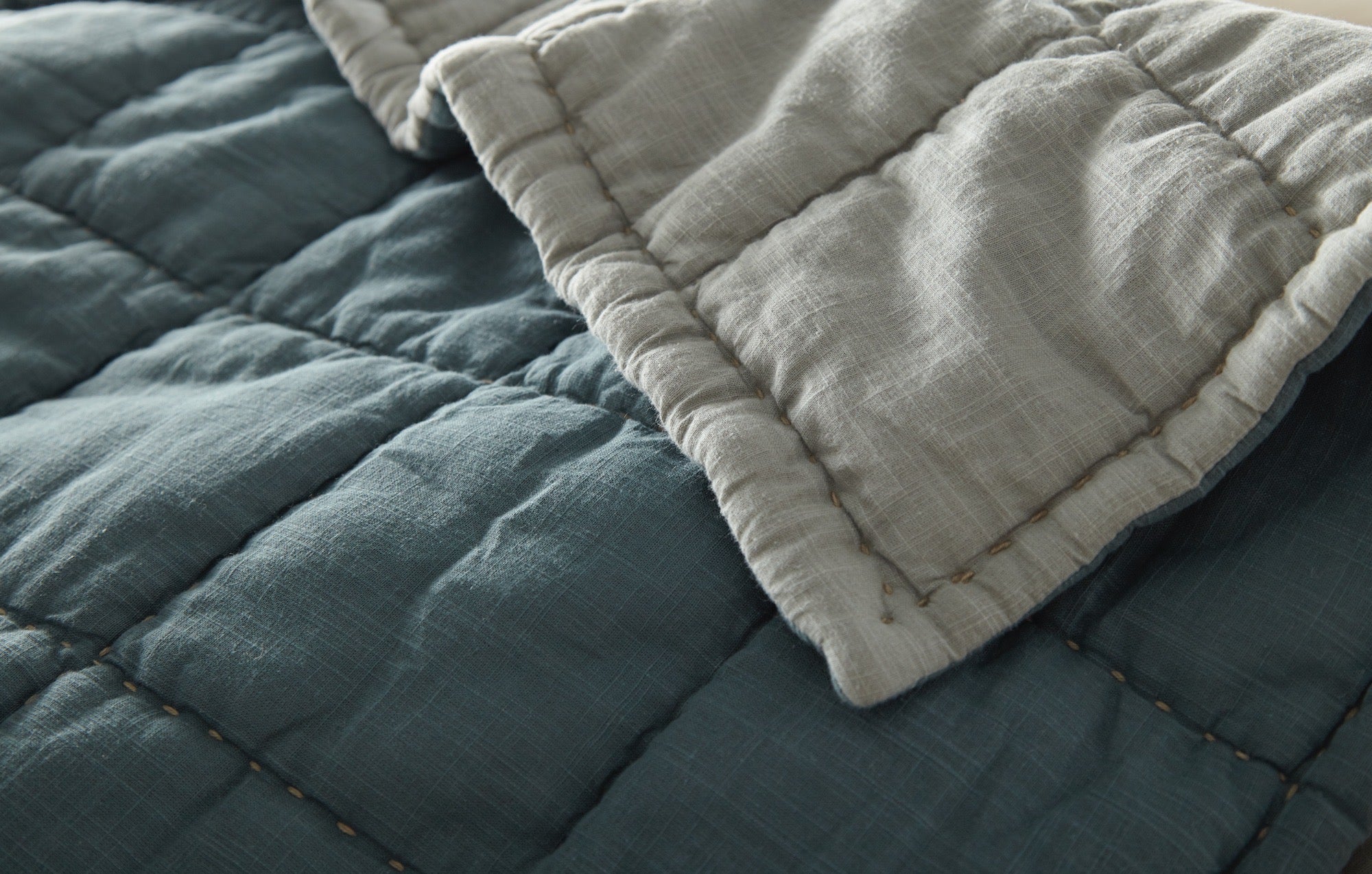 A close-up of the Amity Home Spencer Quilt shows reversible blue slub cotton on one side and light gray on the other, highlighted by visible stitched seams.