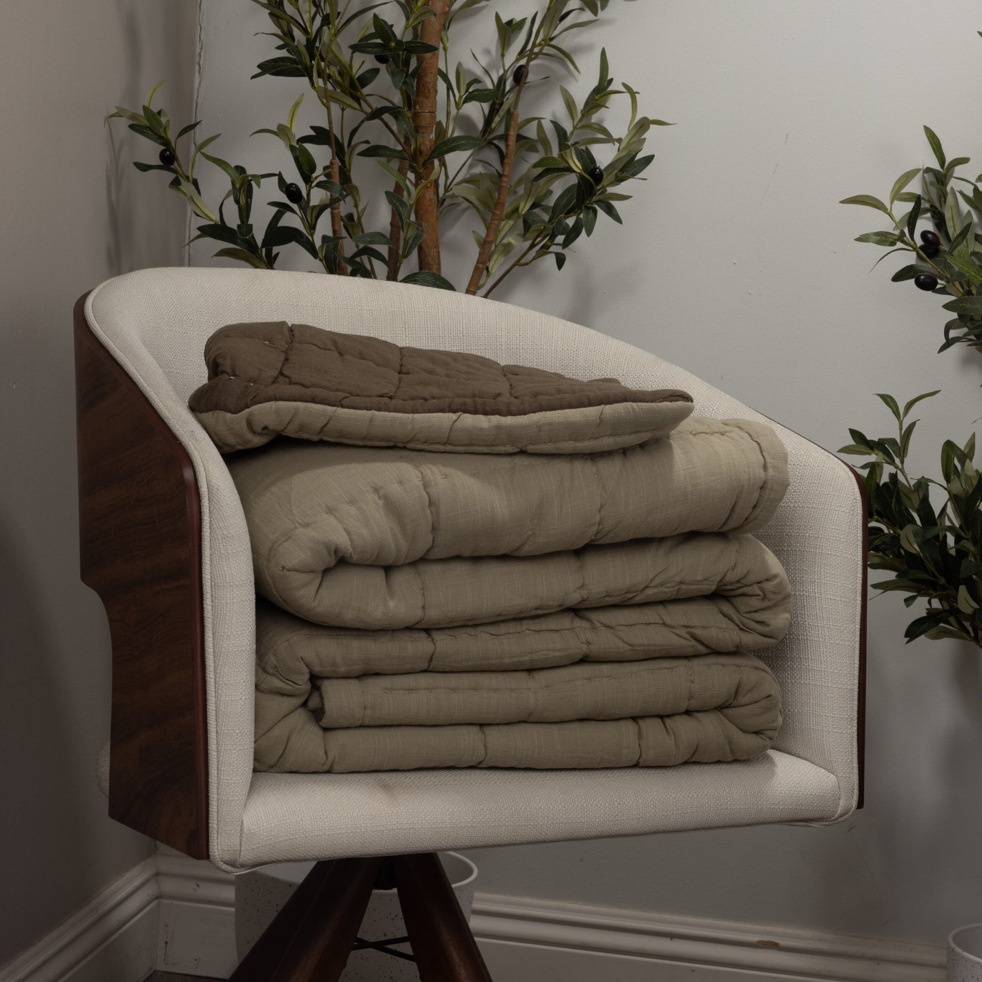 A modern upholstered chair displays three folded Amity Home Spencer Quilts in beige and brown, with green leafy plants set against a light gray wall.