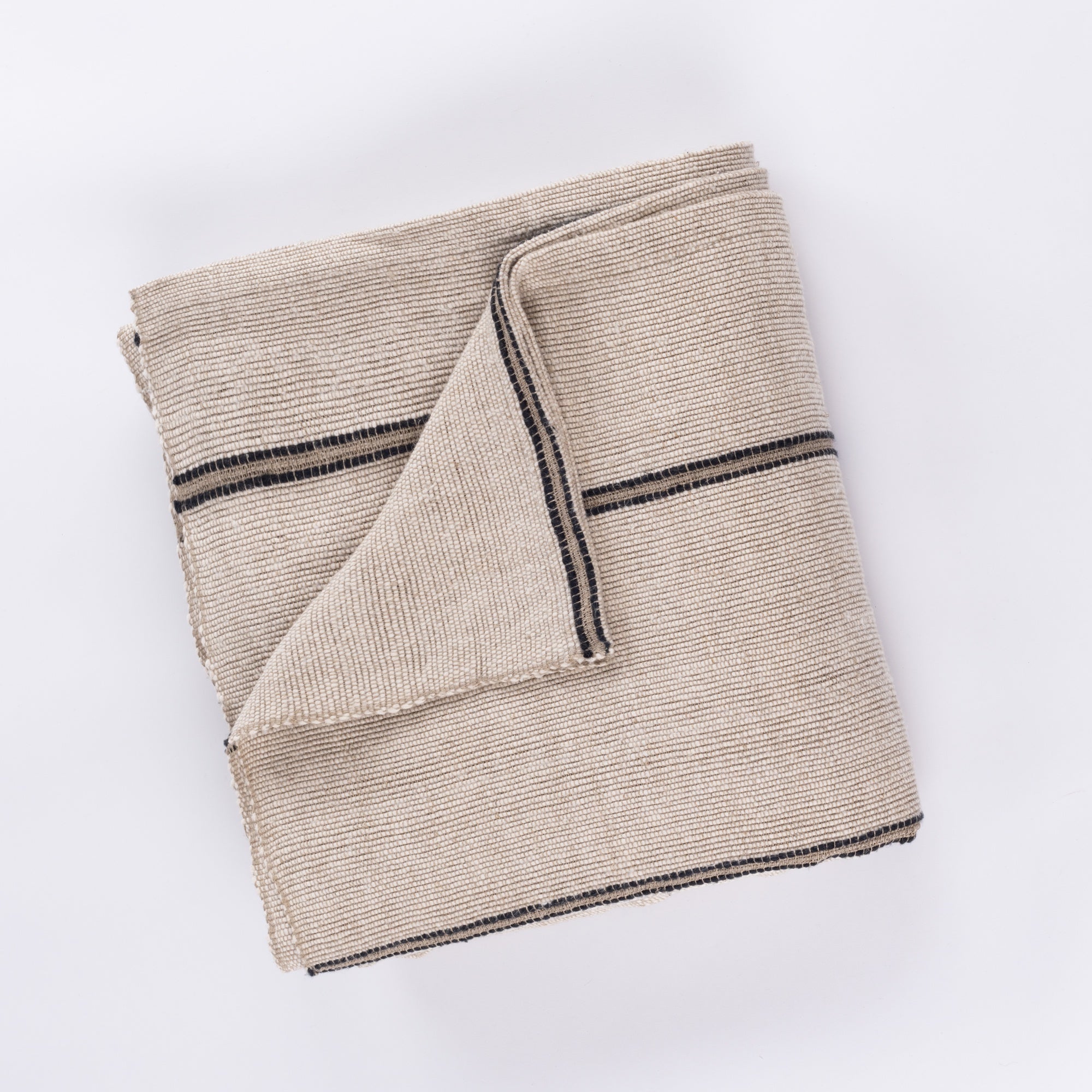 The Rhodes Coverlet Oversized King by Amity Home features a neatly folded beige textured fabric with thin black stripes on white, one corner flipped to reveal the reverse side and its subtle wabi-sabi charm.
