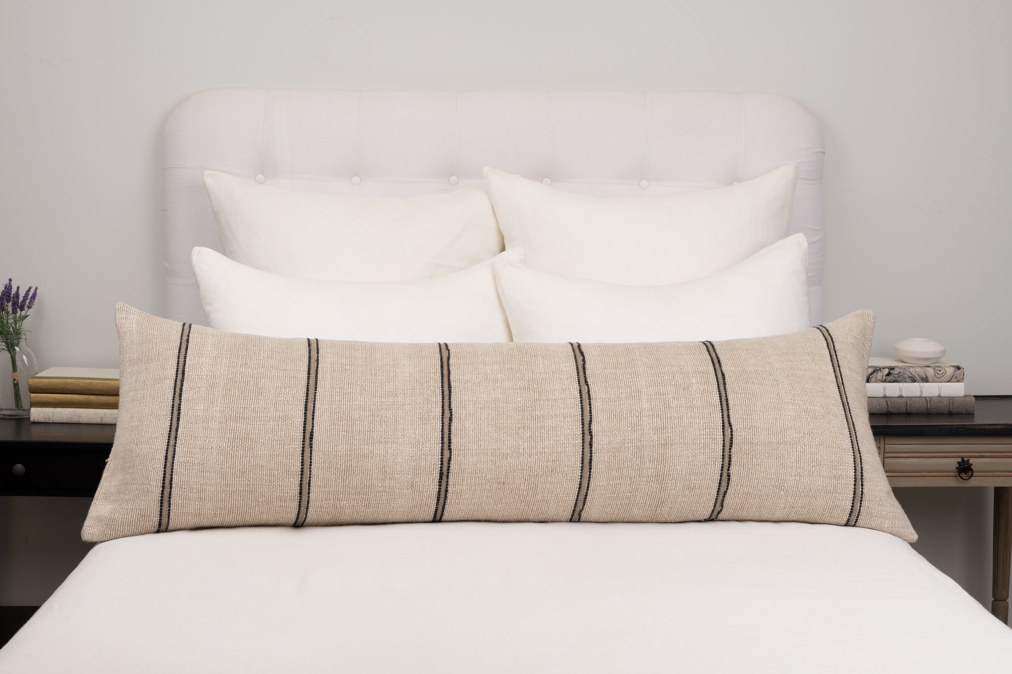 A neatly made bed with a white tufted headboard, four white pillows, and the Amity Home Rhodes Body Pillow Sham—a natural linen bolster with black stripes—sits against a light grey wall. Bedside tables hold books and decor on either side.
