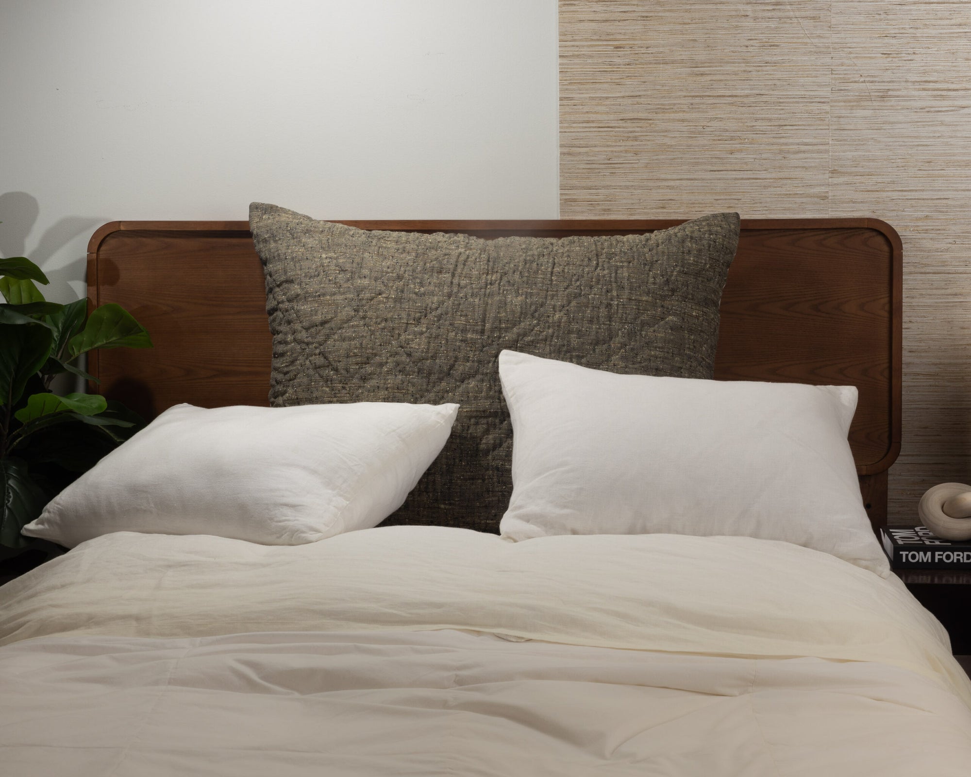 The Adley Dutch Euro by Amity Home features a wooden headboard, white pillows, a large dark textured cushion, and a cream comforter for an organic look. A plant is on the left nightstand; books rest on the right.