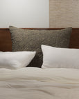 The Adley Dutch Euro by Amity Home features a wooden headboard, white pillows, a large dark textured cushion, and a cream comforter for an organic look. A plant is on the left nightstand; books rest on the right.