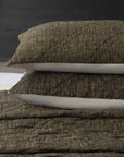 Two Adley Dutch Euro pillows by Amity Home, featuring a brown textured finish for an organic look, are stacked on a bed with a matching blanket against a dark wood headboard and white mattress edge in the background.