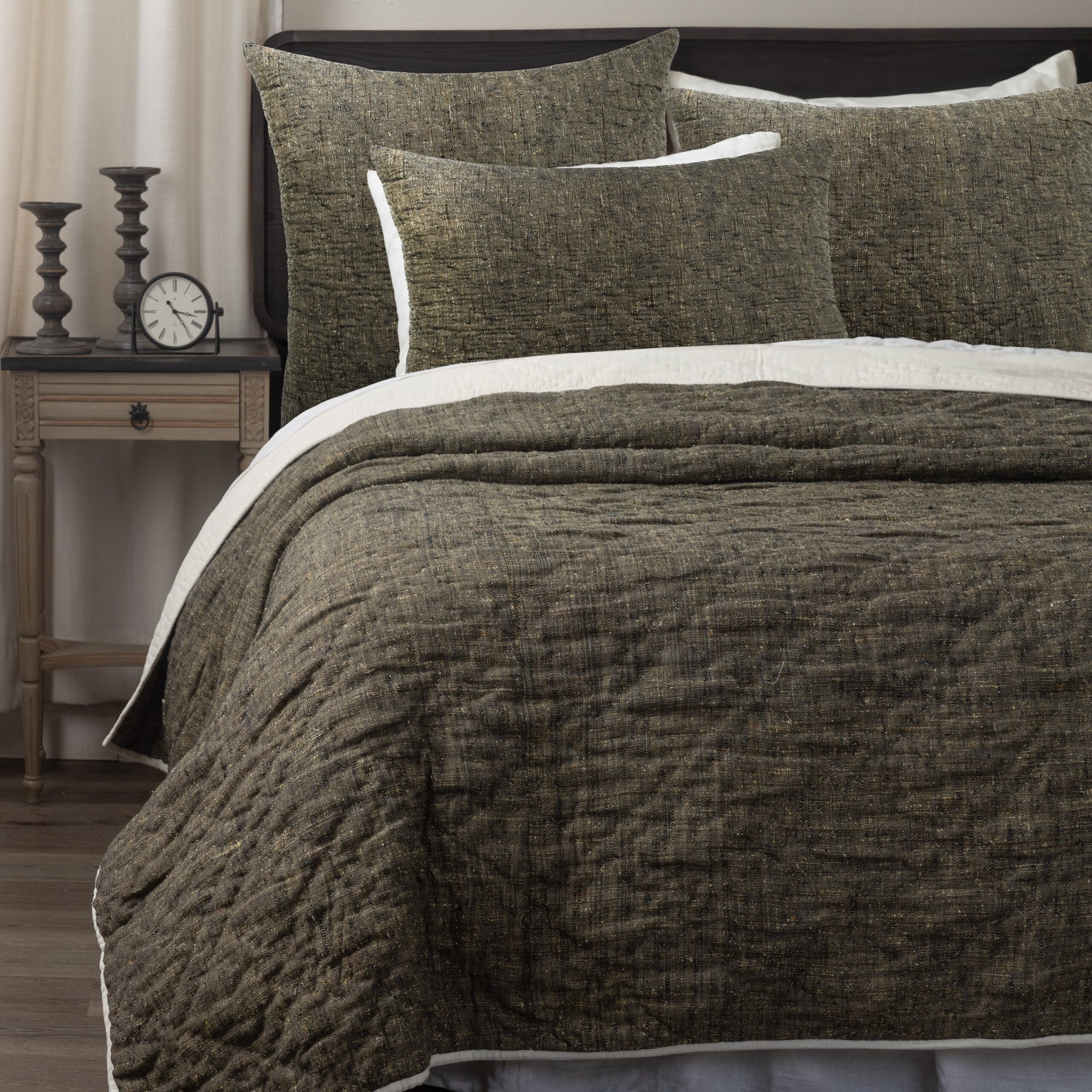 A neatly made bed features the Amity Home Adley Quilt Oversize K—a textured, dark brown, hand-quilted comforter with matching pillows. Next to it stands a bedside table with two candlesticks, a small clock, and light curtains in the background.