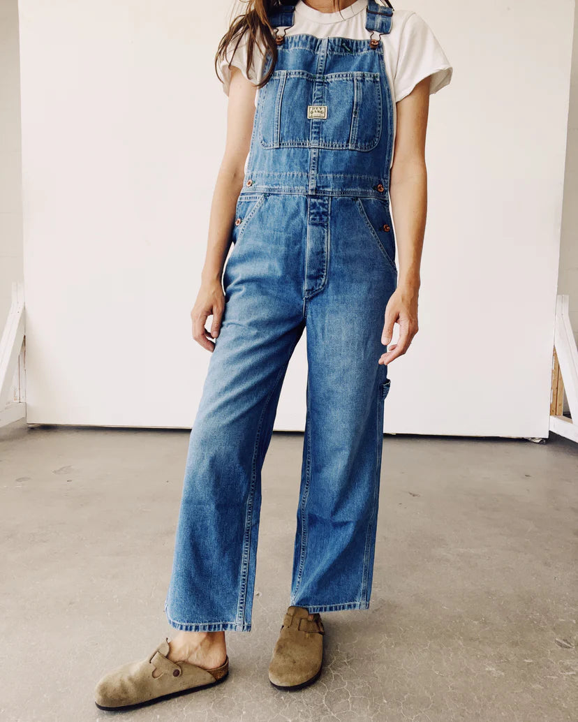 A person stands against a plain background wearing The Miner Overalls by Hey Gang, featuring a unisex fit in vintage washed denim, paired with a white T-shirt. They have on brown casual shoes, with their hands by their sides.