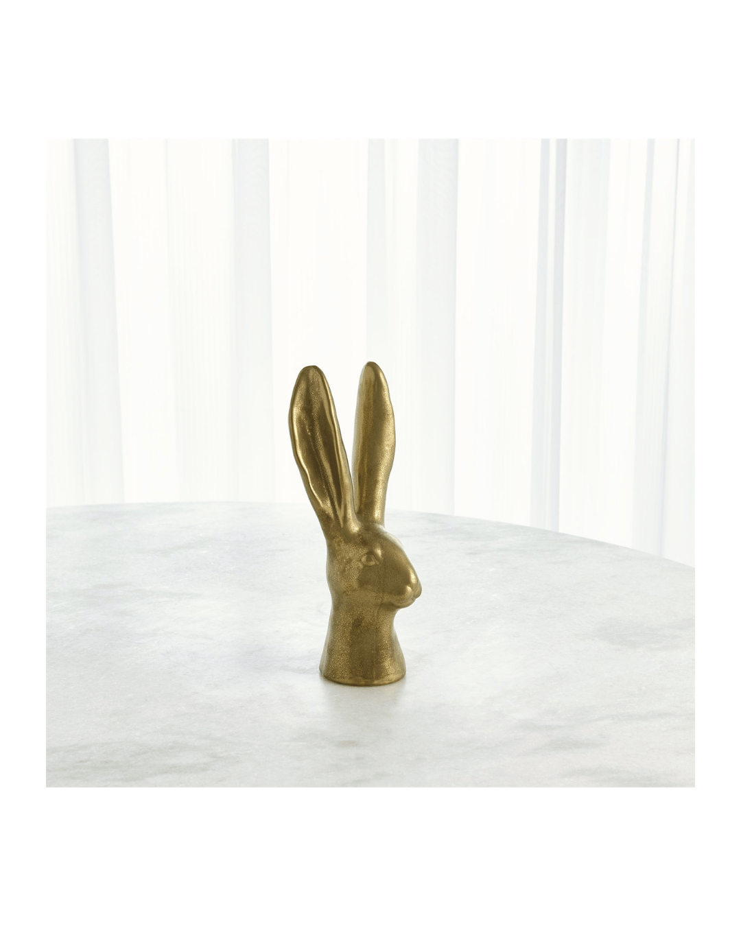 The Rabbit-Matte Gold Small by Global Views, featuring elongated ears, sits on a white marble surface with white drapes in the background, making it a true conversational piece.