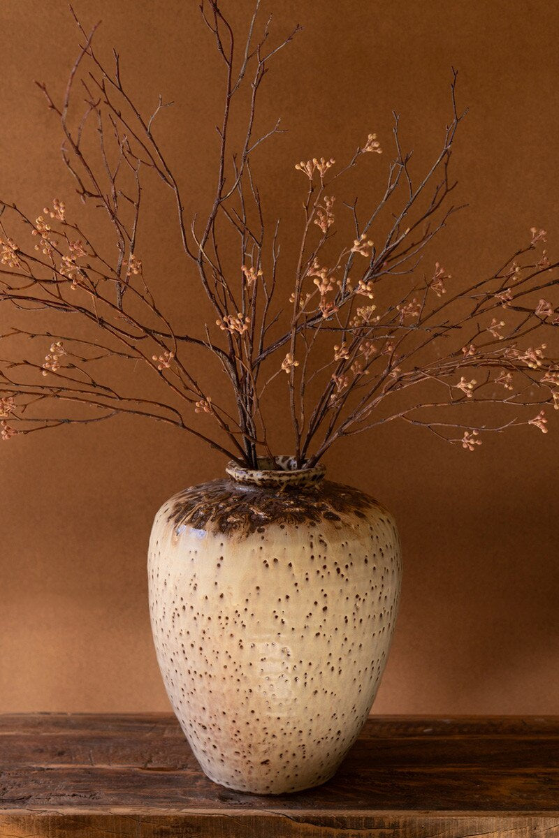 The Large Rustic Ceramic Urn by Kalalou, Inc features a mottled beige and brown finish and holds bare branches with dried buds, set on a wooden surface—an ideal striking entryway accent.