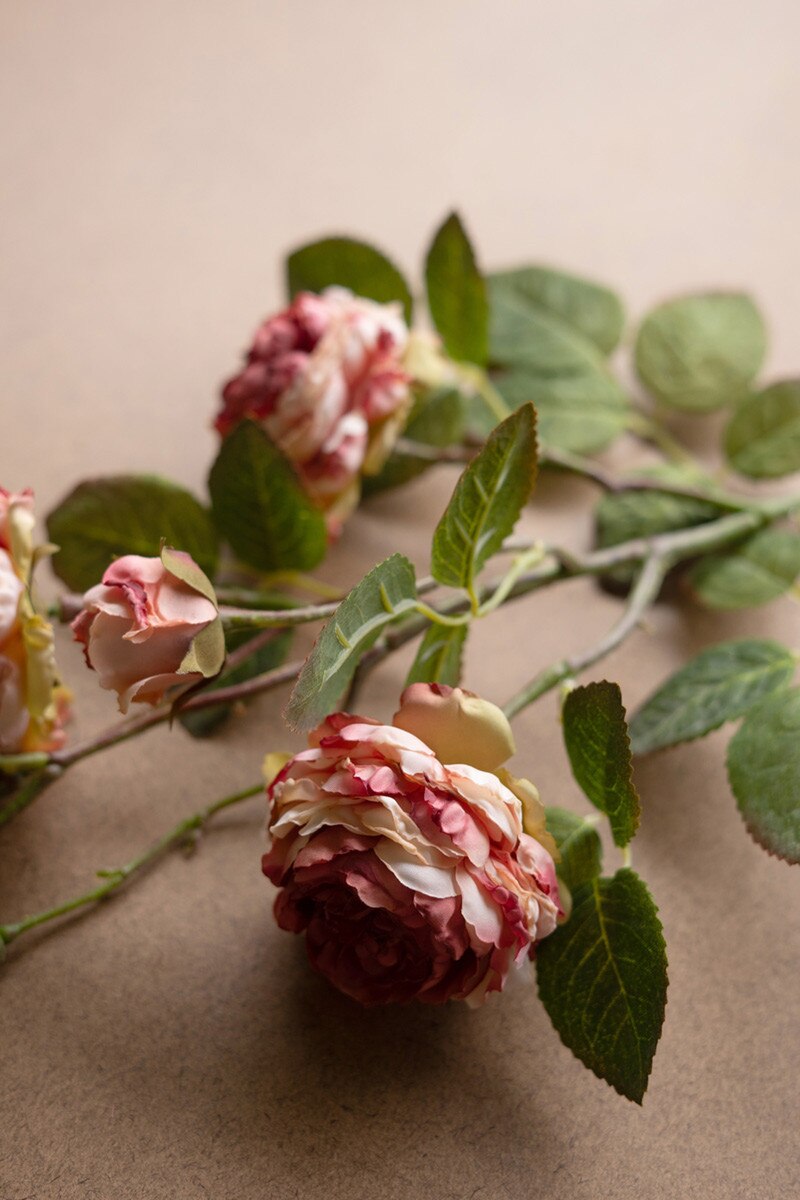 A close-up of three pink and cream Kalalou, Inc Garden Rose artificial flower stems with green leaves on a beige surface—ideal for spring arrangements.