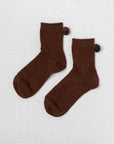 Unified Winter Pom Pom Socks—a cozy brown ankle pair featuring matching side pompoms—are displayed on a lightly textured background.