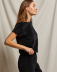 Wearing the Clara Velour Crew Neck Tee from Perfectwhitetee, a person in a black outfit stands in profile against a neutral fabric backdrop. Their long, brown hair cascades down as they look forward with a slight smile. The relaxed fit of the tee adds an effortless elegance, while their left hand rests on their hip with confidence.