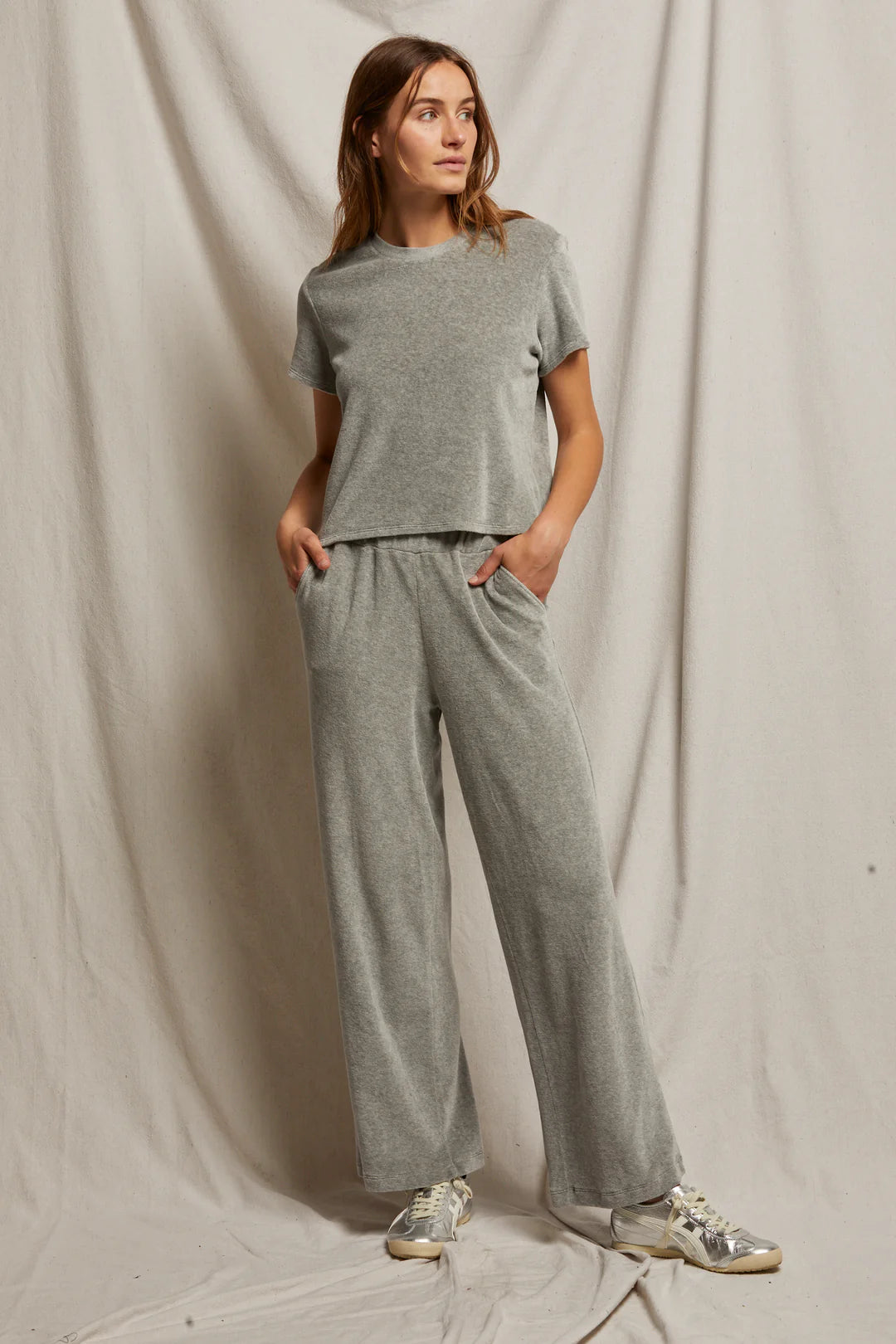 A person stands against a beige fabric backdrop, embodying an effortless style perfect for the holiday season in a Clara Velour Crew Neck Tee by Perfectwhitetee, paired with wide-leg pants. Their hands rest in pockets, sneakers adorn their feet, and their hair flows loosely as they gaze off to the side.