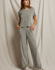 A person stands against a beige fabric backdrop, embodying an effortless style perfect for the holiday season in a Clara Velour Crew Neck Tee by Perfectwhitetee, paired with wide-leg pants. Their hands rest in pockets, sneakers adorn their feet, and their hair flows loosely as they gaze off to the side.