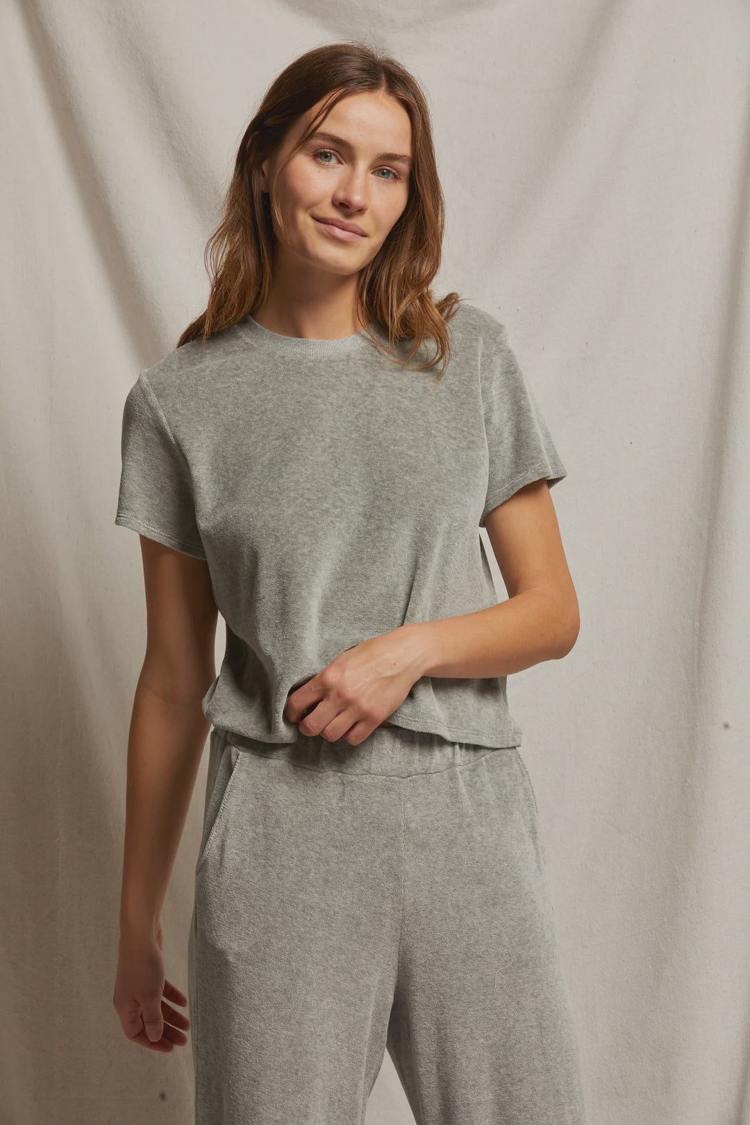 A woman wearing the Clara Velour Crew Neck Tee by Perfectwhitetee, styled casually in light grey with matching pants that create a boxy silhouette, stands against a plain fabric backdrop. She gazes at the camera with a slight smile, her long, wavy hair cascading loosely.