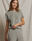 A woman wearing the Clara Velour Crew Neck Tee by Perfectwhitetee, styled casually in light grey with matching pants that create a boxy silhouette, stands against a plain fabric backdrop. She gazes at the camera with a slight smile, her long, wavy hair cascading loosely.