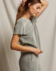 A person with long hair is wearing a Clara Velour Crew Neck Tee in grey, complemented by matching pants, standing sideways with one hand touching the back of their head. This ensemble from Perfectwhitetee, featuring a boxy silhouette, evokes a sense of cozy elegance against a plain, light-colored fabric backdrop.