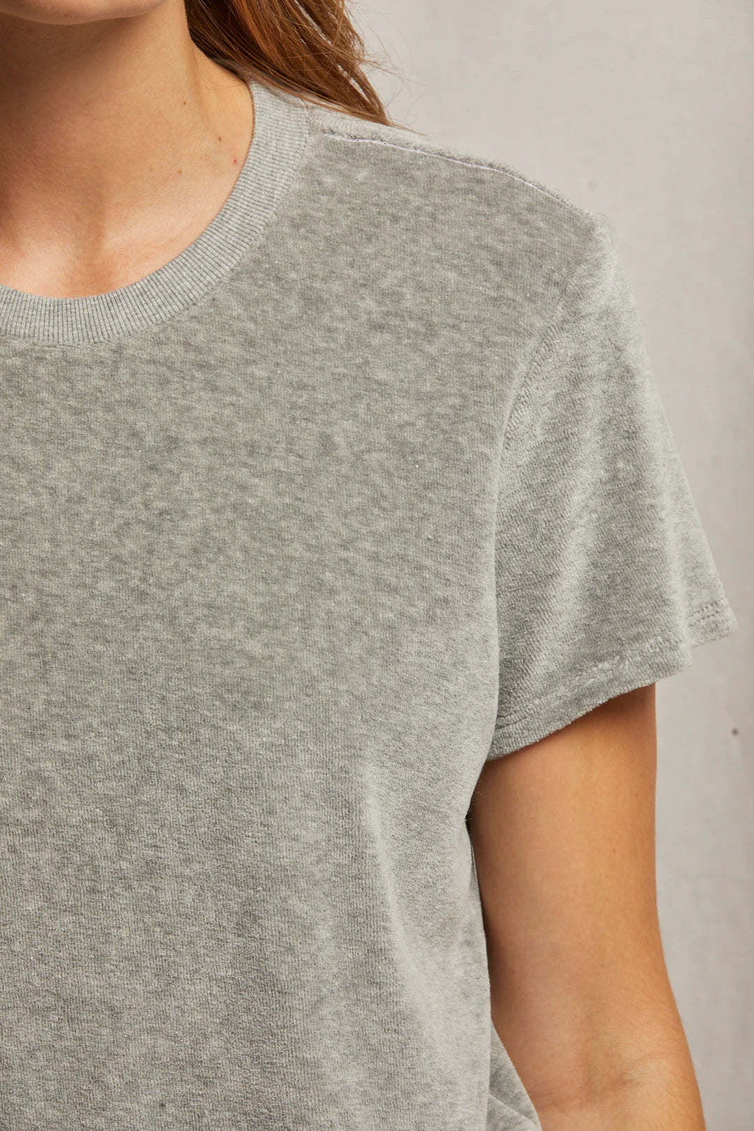 A person is wearing the Clara Velour Crew Neck Tee by Perfectwhitetee, a light gray, short-sleeve shirt with a boxy silhouette. The image features the upper half of the torso and part of the left arm against a neutral backdrop, highlighting the subtly textured material that exudes casual comfort ideal for the holiday season.