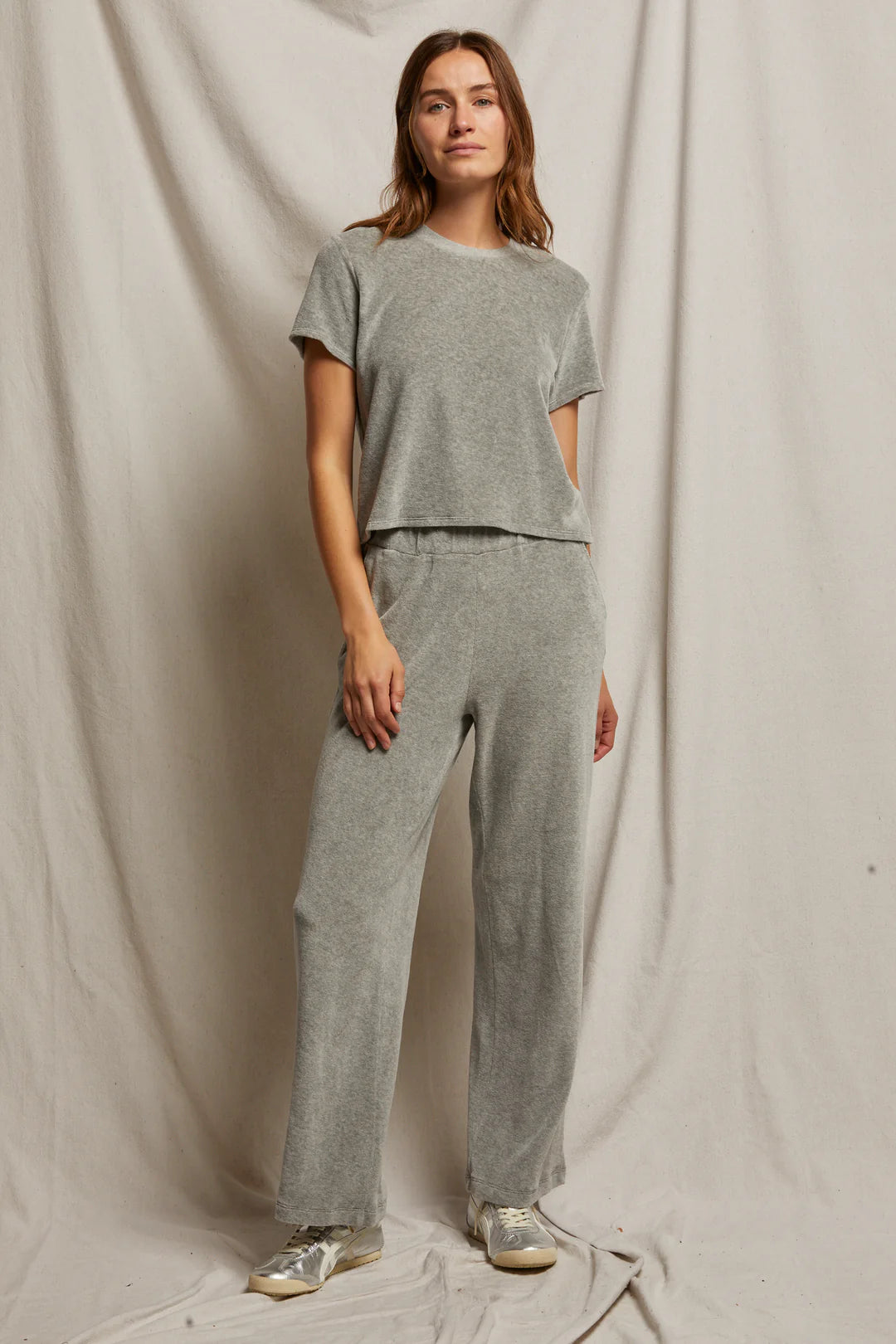 A person stands against a draped fabric background, wearing a light gray short-sleeve top and the Holly Velour Wide Leg Sweatpant by Perfectwhitetee. The pants are crafted from plush velour fabric, and the look is completed with light-colored sneakers, one hand casually in a pocket for a relaxed posture.