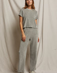 A person stands against a draped fabric background, wearing a light gray short-sleeve top and the Holly Velour Wide Leg Sweatpant by Perfectwhitetee. The pants are crafted from plush velour fabric, and the look is completed with light-colored sneakers, one hand casually in a pocket for a relaxed posture.