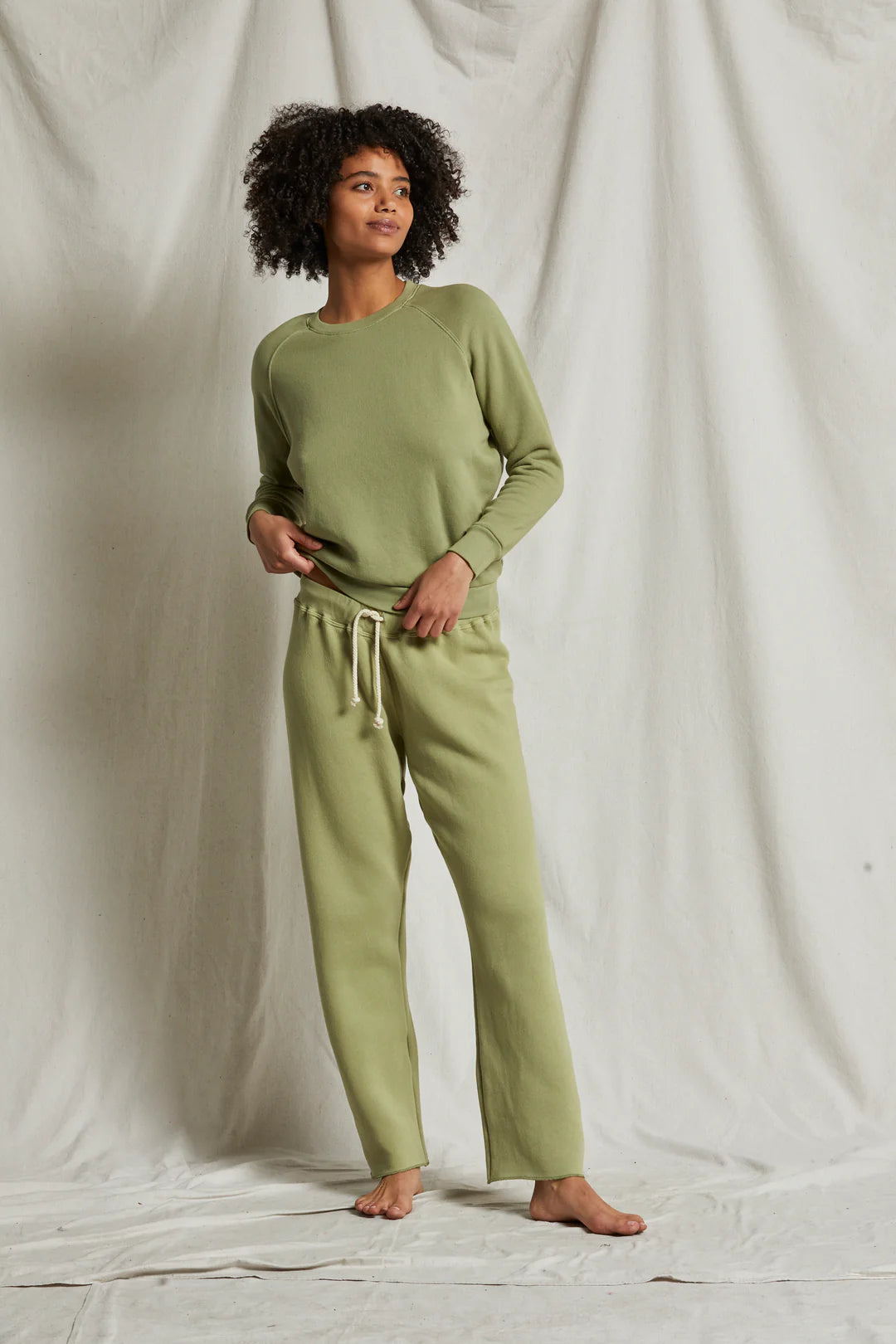 A person with curly hair stands barefoot on a white fabric backdrop. They are wearing a sage green long-sleeve shirt and the matching Collins Fleece Straight Leg Sweatpant by Perfectwhitetee, featuring a drawstring waist that creates a casual sweatpant silhouette. The individual looks slightly to the side with a relaxed posture and a subtle smile.