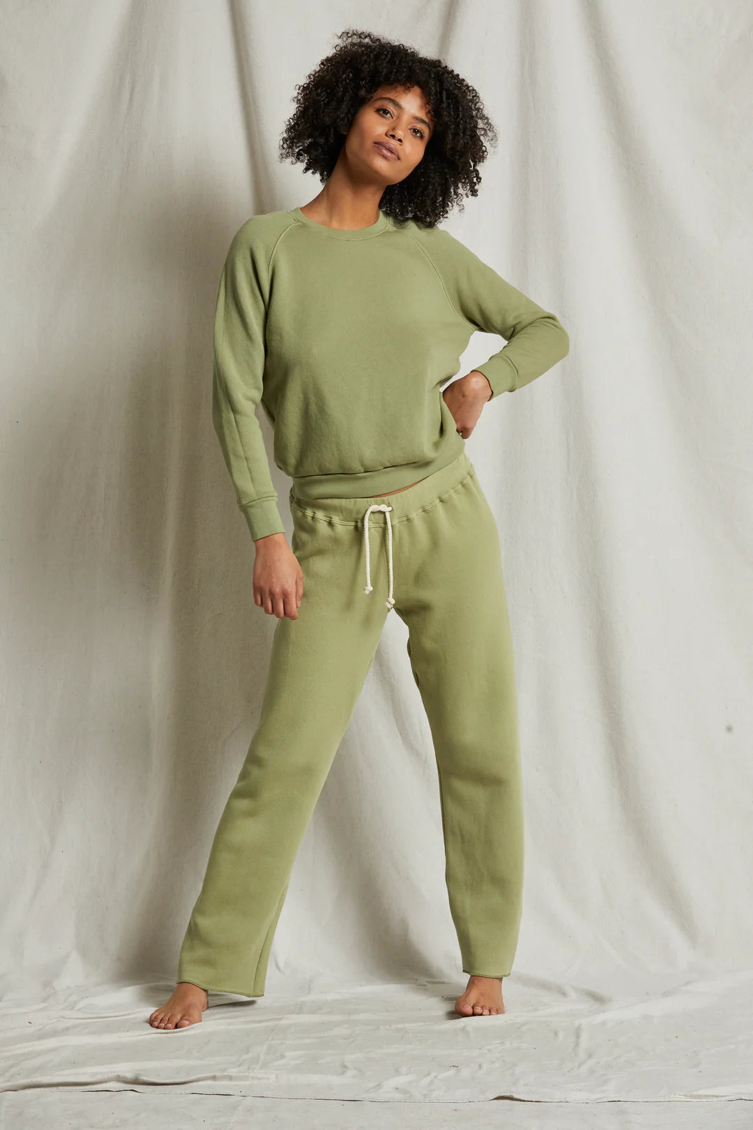 A person with curly hair stands against a light grey fabric backdrop, wearing a matching light green Perfectwhitetee sweatshirt and Collins Fleece Straight Leg Sweatpant. The relaxed fit of their cotton fleece outfit complements the straight-leg silhouette of the sweatpants. Their left hand is on their hip, and they are barefoot.