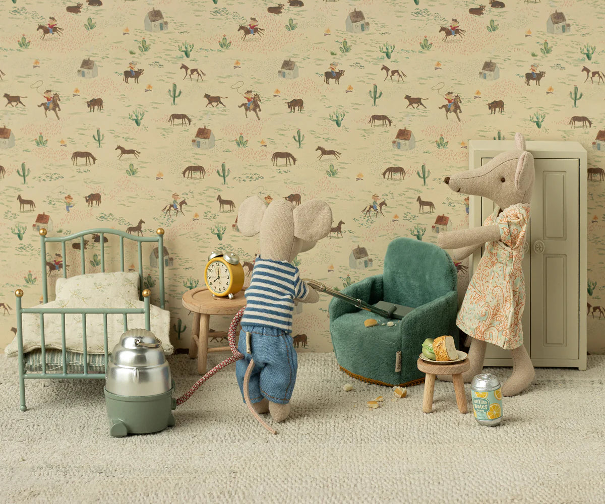 Two Maileg toy mice in a miniature room: one vacuums by a Maileg Folding Chair, while the other mouse in a dress stands nearby. The cozy Mouse hole Farmhouse features a bed, clock, crumbs, and playful animal-themed wallpaper.