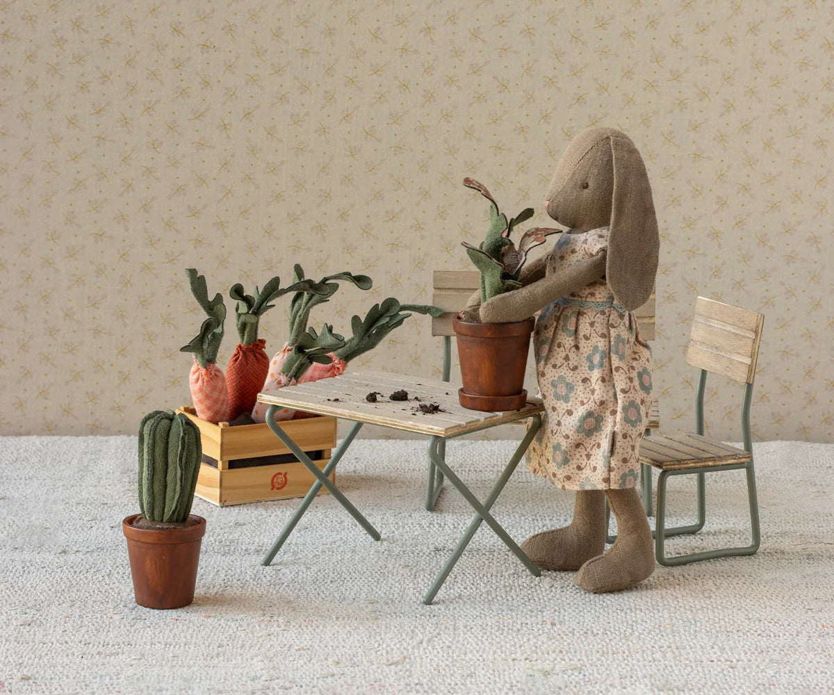 A Maileg bunny in a floral dress stands by a small table, potting the Maileg Flower Pot. Nearby are a wooden crate with fabric carrots, two chairs, and a potted fabric cactus on the floor.