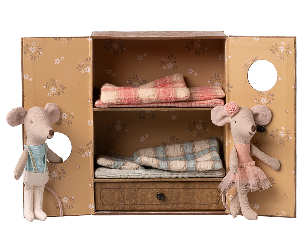 Two Maileg Ballet Mice—Little Sis in a pink tutu and headband, Little Bro in blue overalls—stand by an open toy wardrobe with floral wallpaper, folded plaid blankets, and a drawer. A charming Maileg set for any mouse house or ballet playset.