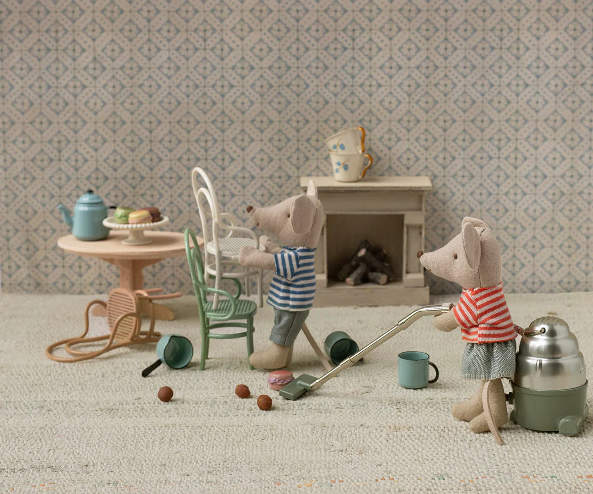 Two Maileg toy mice in striped shirts tidy a cozy room: one vacuums pebbles, the other lifts a Vintage Chair. The dollhouse-like space is filled with charming mice furniture, patterned decor, dishes on a table, and a glowing fireplace.