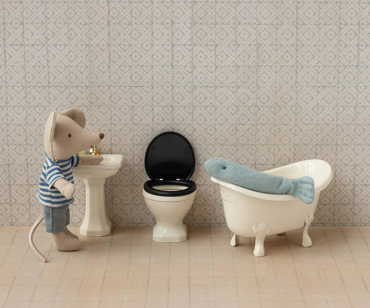 A stuffed mouse in a striped shirt stands at the Maileg Water Closet sink by Maileg, next to a toy toilet and bathtub, where a blue plush seal lies; the scene evokes a cozy small bathroom.