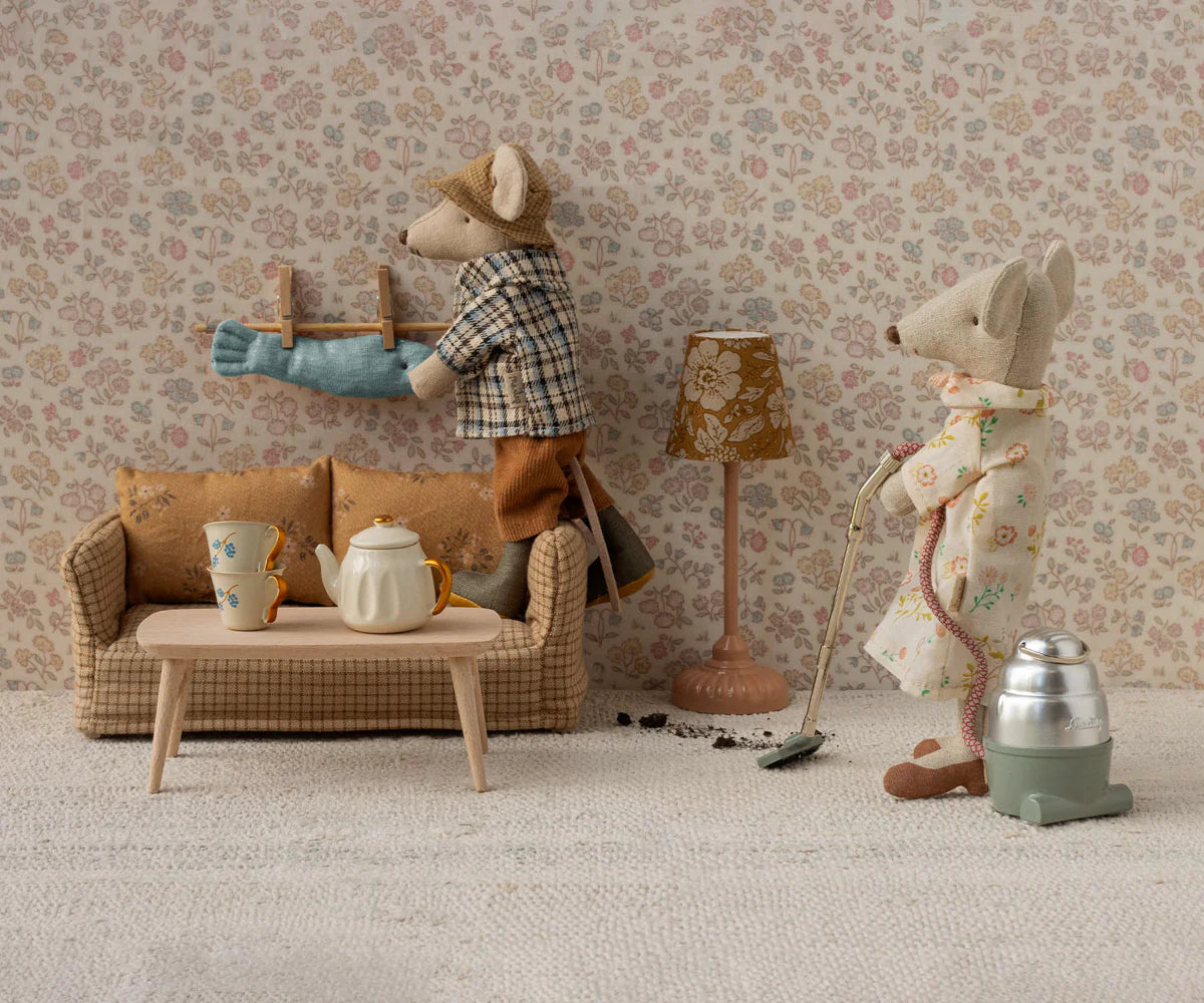 Two mice from Maileg tidy their vintage living room: one vacuums, the other hangs laundry. A soft Maileg Couch, table with teapot and cups, lamp, and patterned wallpaper add to the cozy charm.