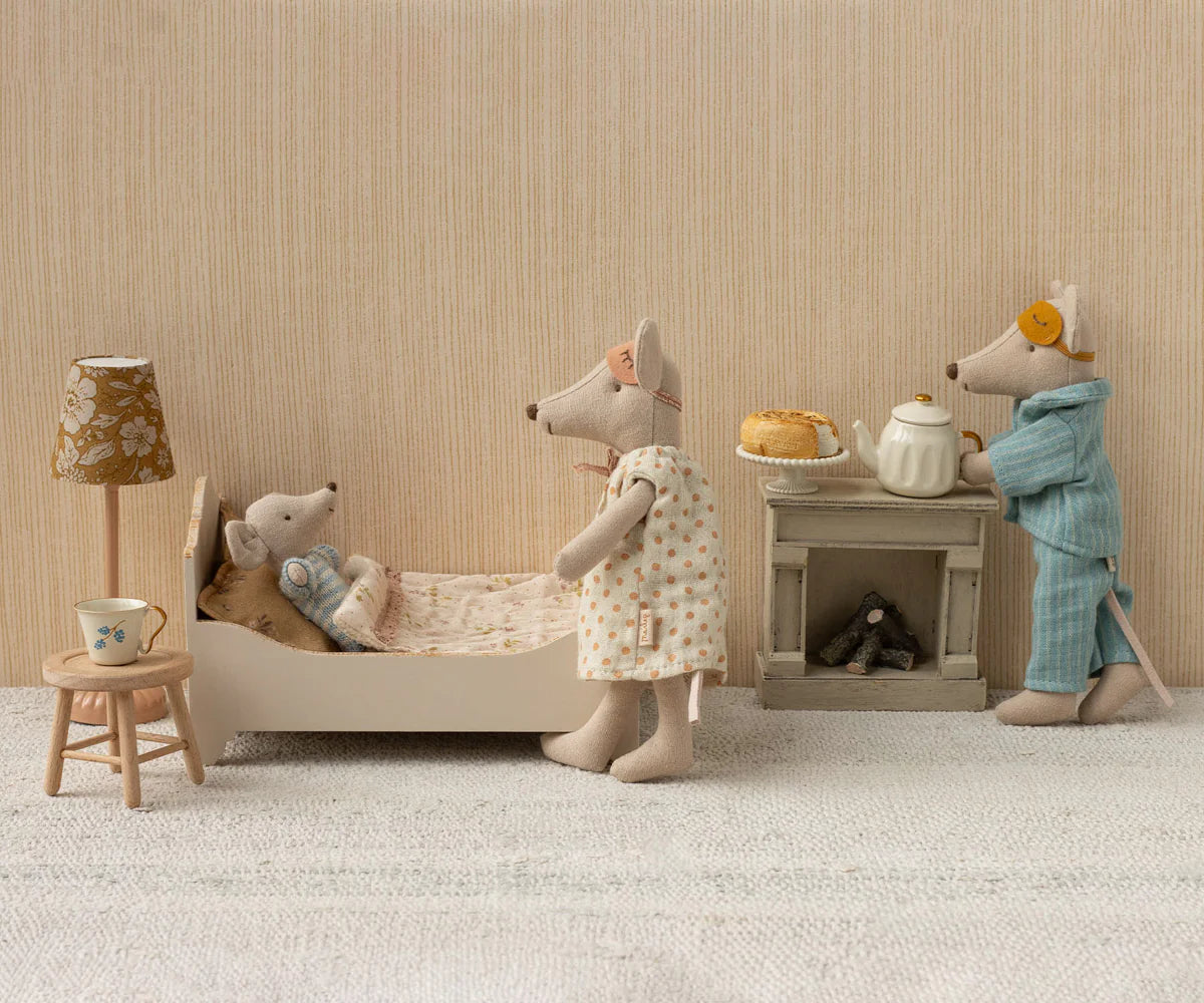 Three Maileg toy mice enjoy a cozy room: a baby sleeps in the Maileg Bed with soft linens, while an adult mouse in a dress stands by and another in pajamas with a sleep mask makes tea. A lamp and stool with cup complete the scene.