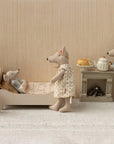 Three Maileg toy mice enjoy a cozy room: a baby sleeps in the Maileg Bed with soft linens, while an adult mouse in a dress stands by and another in pajamas with a sleep mask makes tea. A lamp and stool with cup complete the scene.