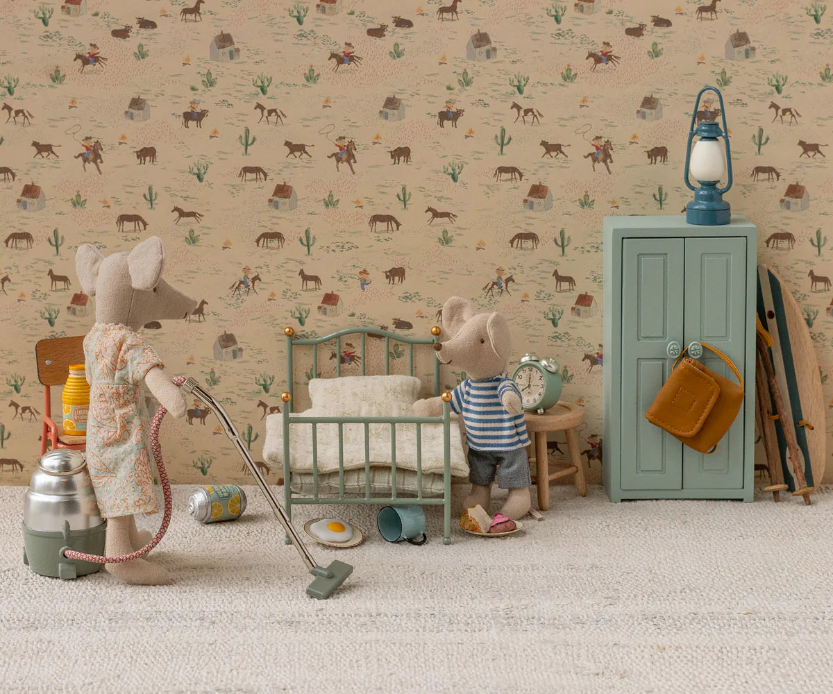 Two toy mice in a miniature room: one vacuums while the other stands by a Maileg Vintage Single Bed. Scattered toys, a green wardrobe, lamp, and western animal-and-cacti wallpaper complete the charming Maileg scene.