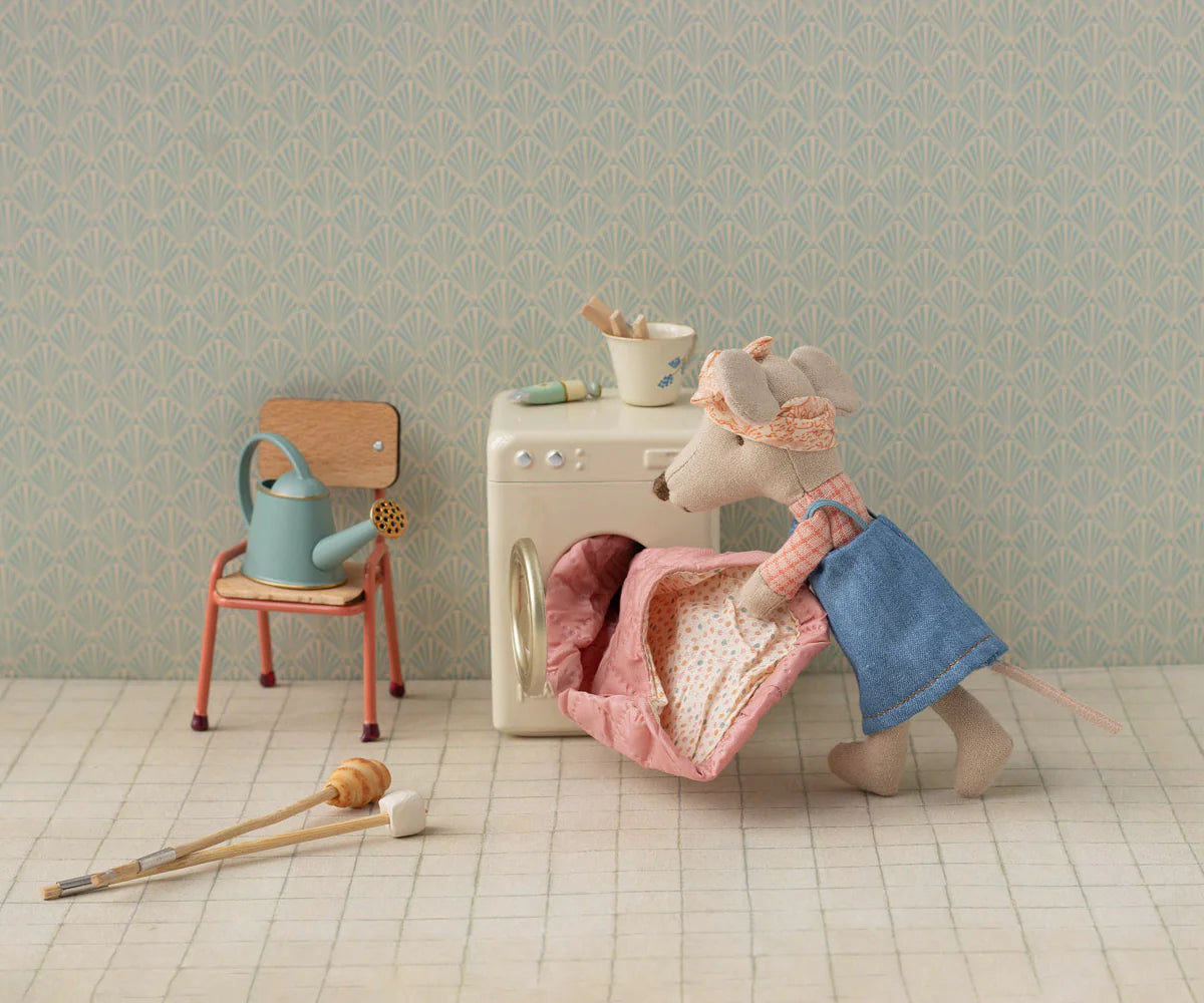 The Maileg Hiker Mouse, dressed in a blue outfit and headband, pulls a pink quilt from a toy washing machine. Nearby are fun accessories: a watering can on a chair, bowl on the machine, and mop on the tiled floor. Brand: Maileg.