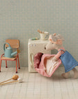 The Maileg Hiker Mouse, dressed in a blue outfit and headband, pulls a pink quilt from a toy washing machine. Nearby are fun accessories: a watering can on a chair, bowl on the machine, and mop on the tiled floor. Brand: Maileg.
