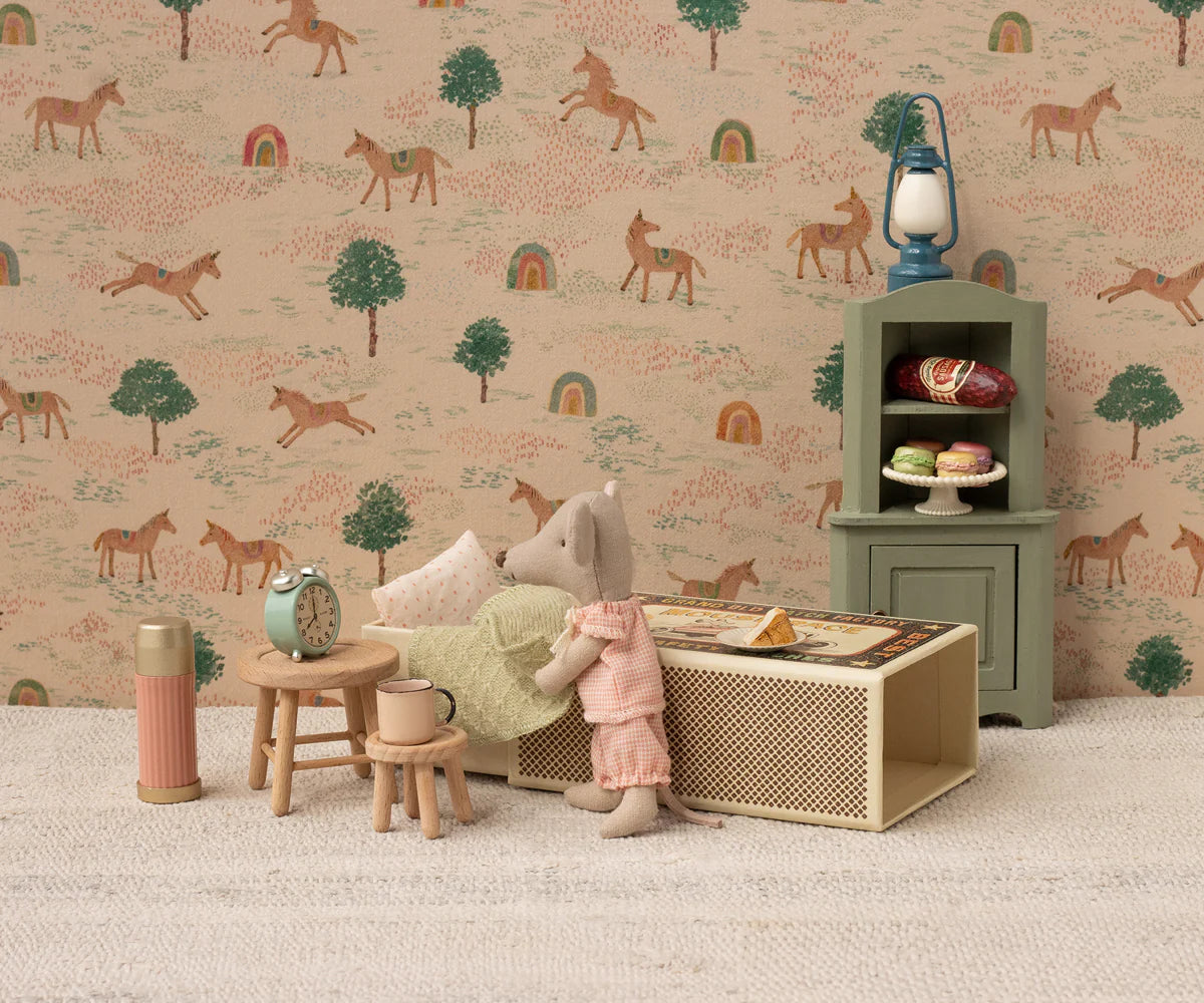The Maileg Little Sister Mouse / Matchbox by Maileg wears pajamas and stands in a cozy scene with a bed, nightstand, clock, and stool. A backdrop with horses, trees, and rainbows adds a whimsical touch to this charming collectible.