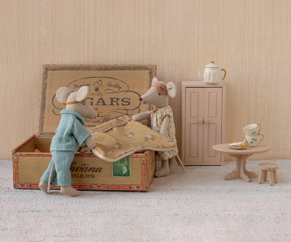 Maileg Mum &amp; Dad Mice in a cigar box bed—one wears pajamas, the other a dress and bow. Nearby are a small cabinet, round table with teapot and cake, and a tiny wooden stool. All by Maileg.